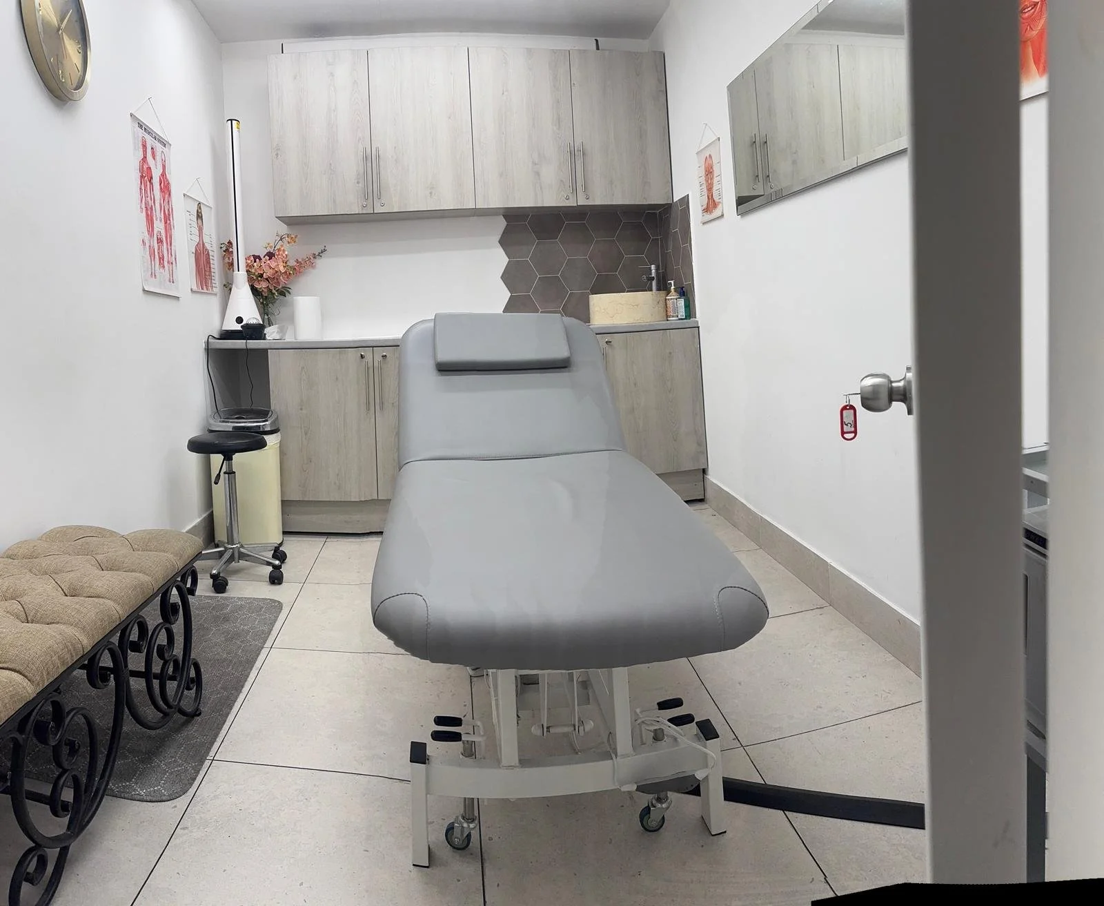 Medical examination room with a treatment bed, medical supplies, decorative wall art, and a small sink area.