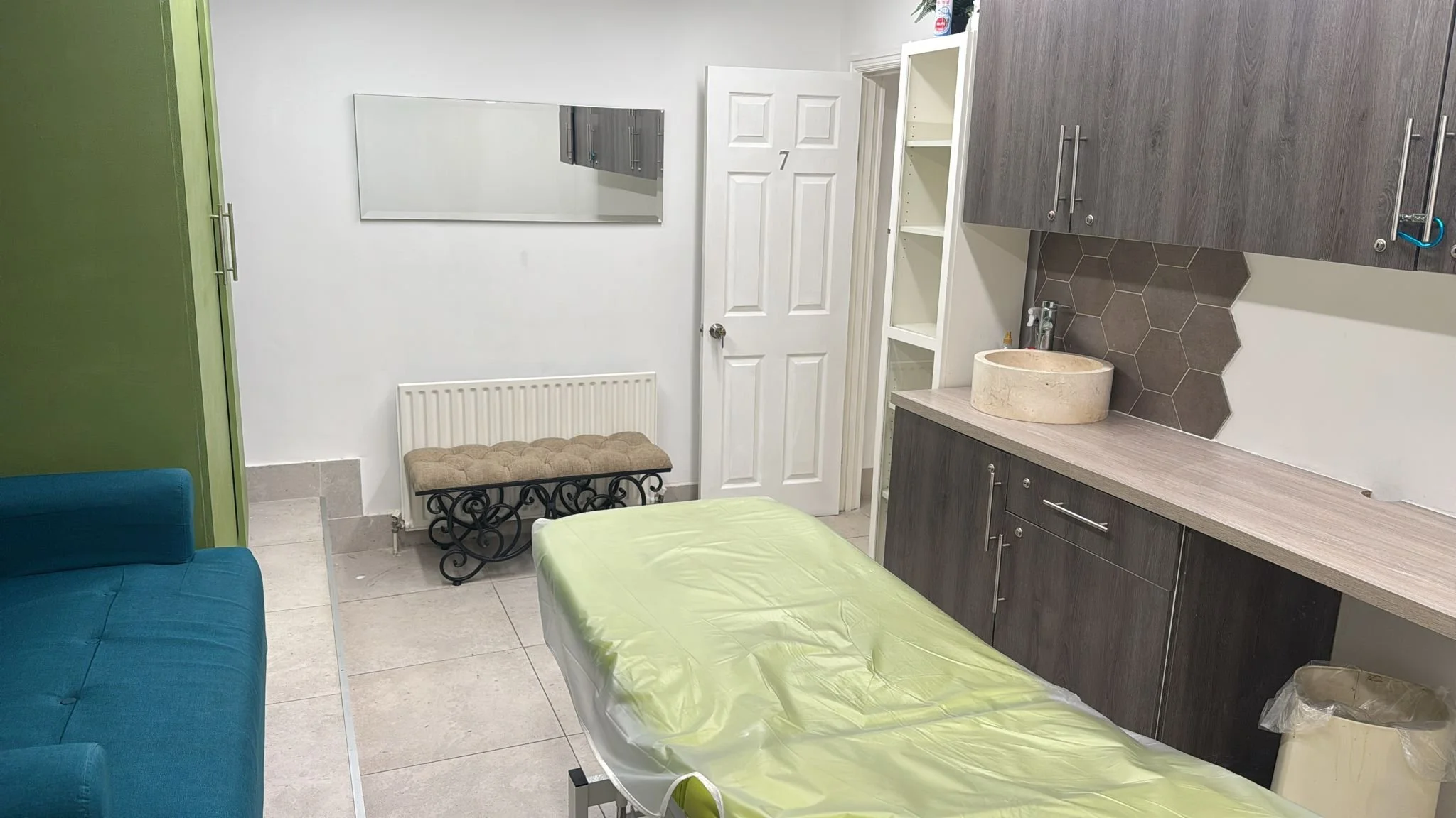 Room with a green and blue couch, a massage table with a yellow sheet, a white door, a mirror, a small bench, a cabinet with a stone sink, and hexagonal tile backsplash.