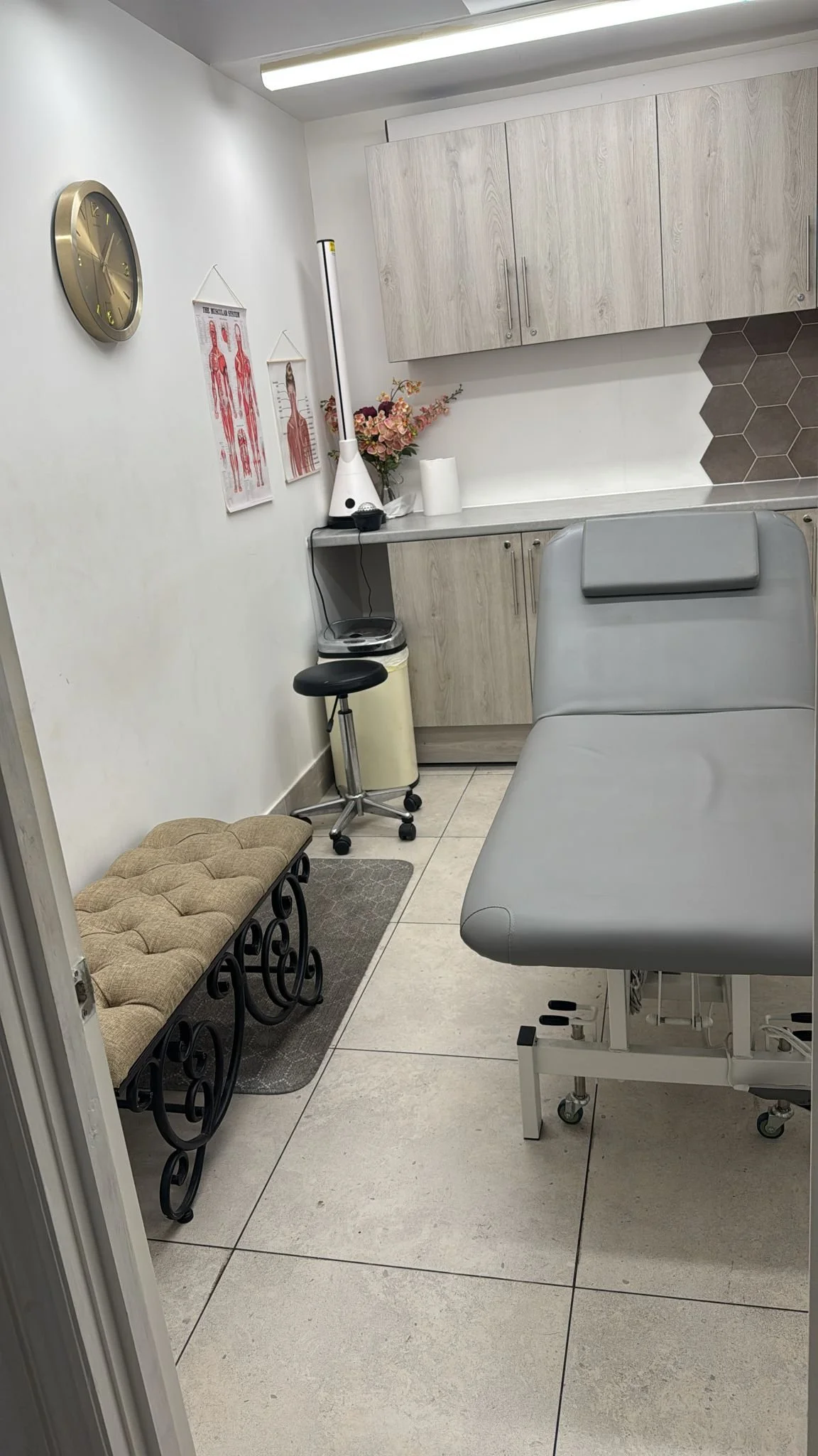 Medical examination room with an examination table, a beige cushioned bench, a stool, medical posters, and cabinets.