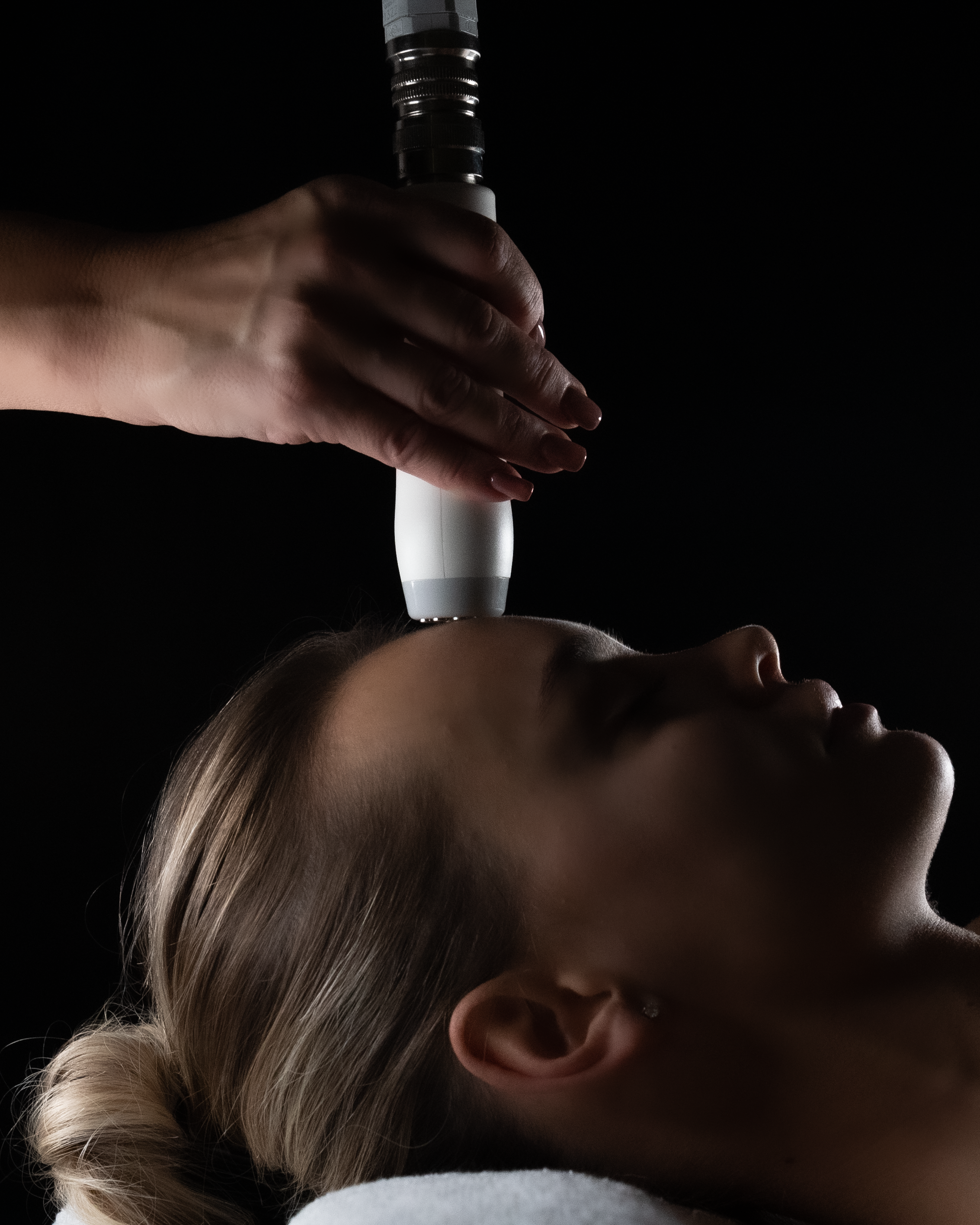A person receiving a facial treatment with a handheld device in a dark, soothing setting.