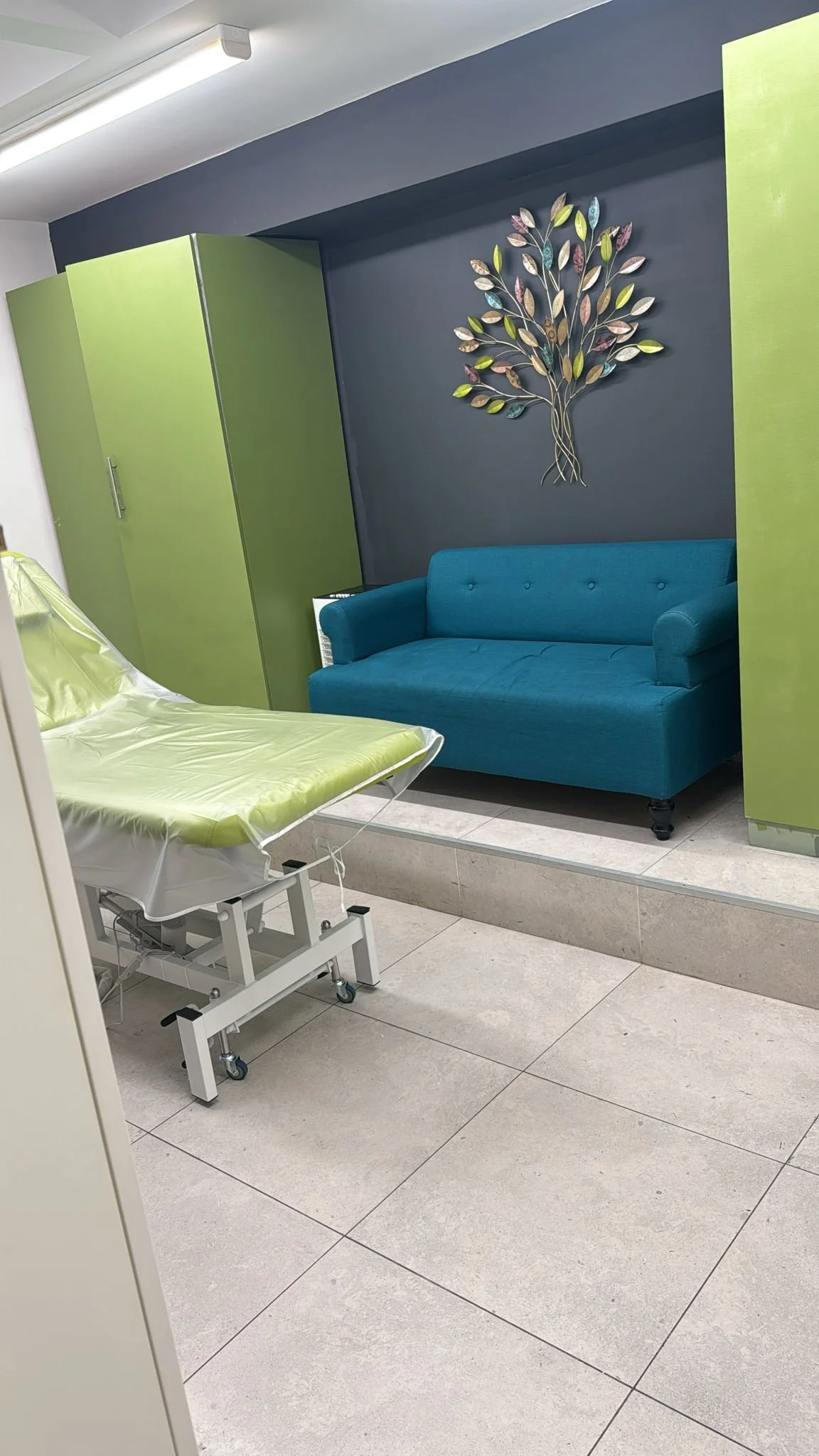 A hospital room with a hospital bed covered with a green sheet, a teal sofa, green cabinets, and a decorative metal tree on a blue-gray wall.