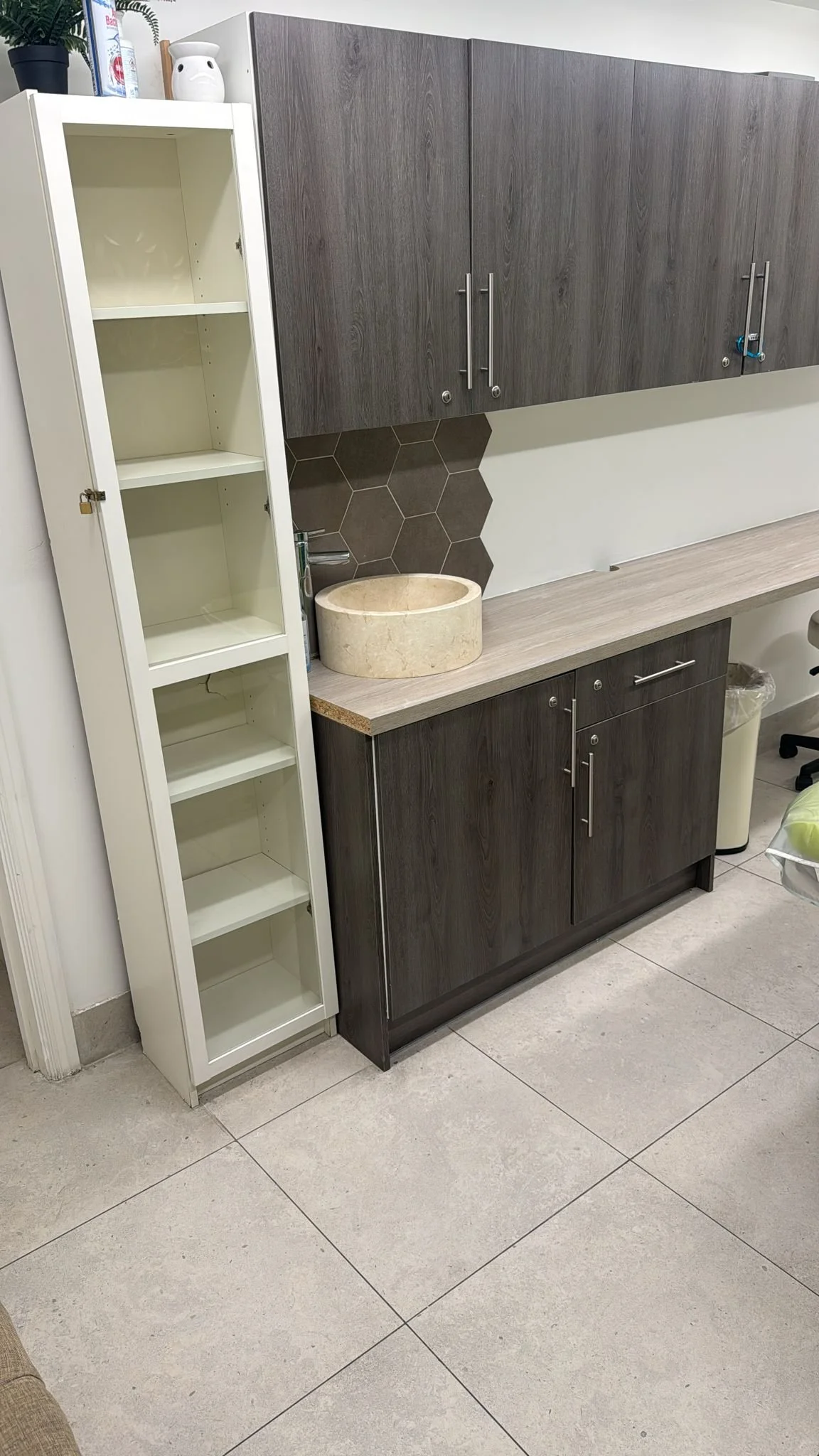 A kitchen corner with a white empty bookcase, dark wood cabinet, hexagonal tiled wall, beige stone sink, and countertop.