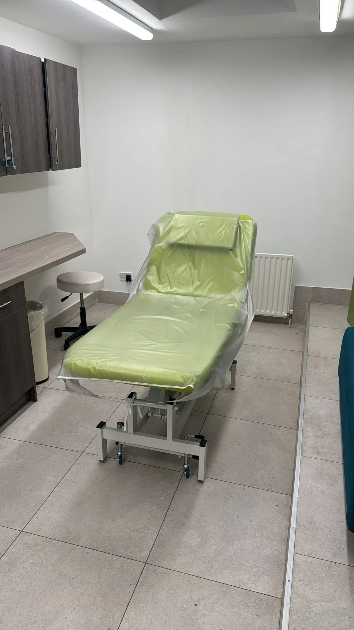 A medical examination room featuring an empty hospital bed with green protective sheets, a padded stool, wall-mounted cabinets, and tiled flooring.