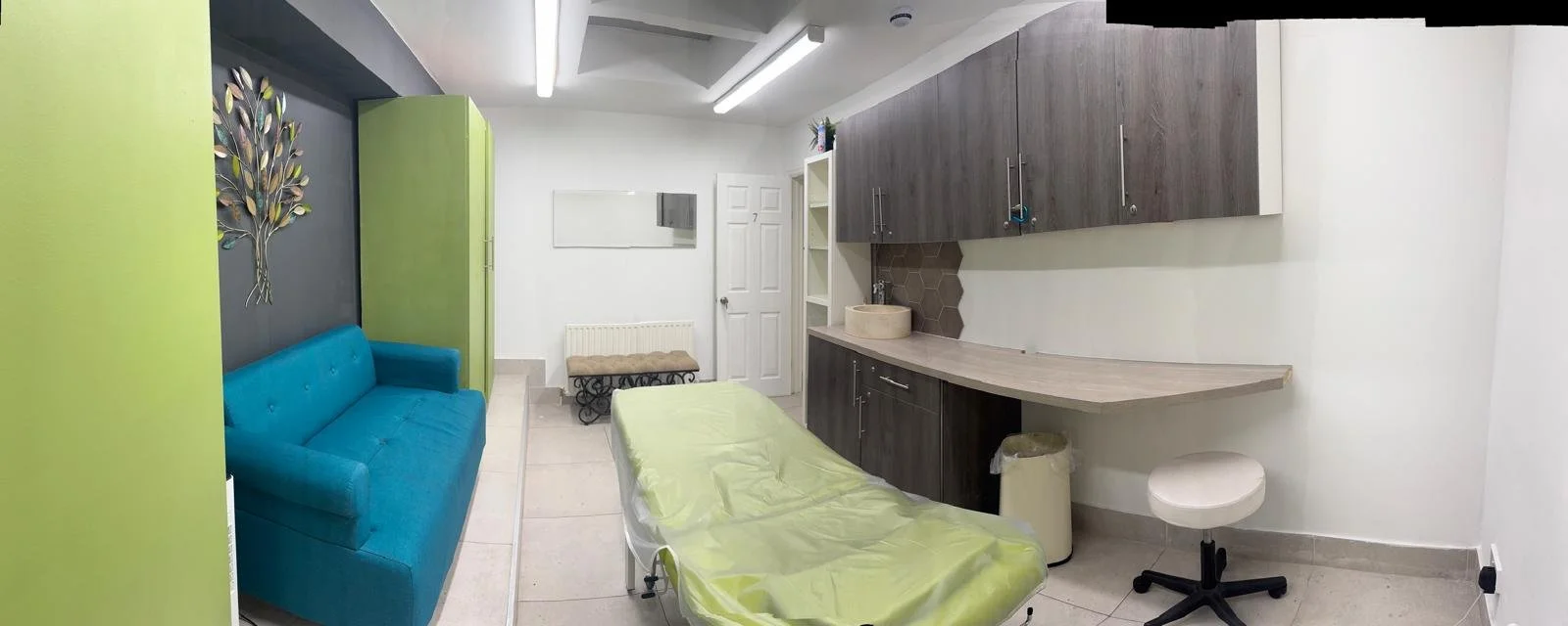 Medical examination room with a blue couch, examination bed covered with a green sheet, dark wood cabinets, a white rolling stool, and a sink area with beige tiles.