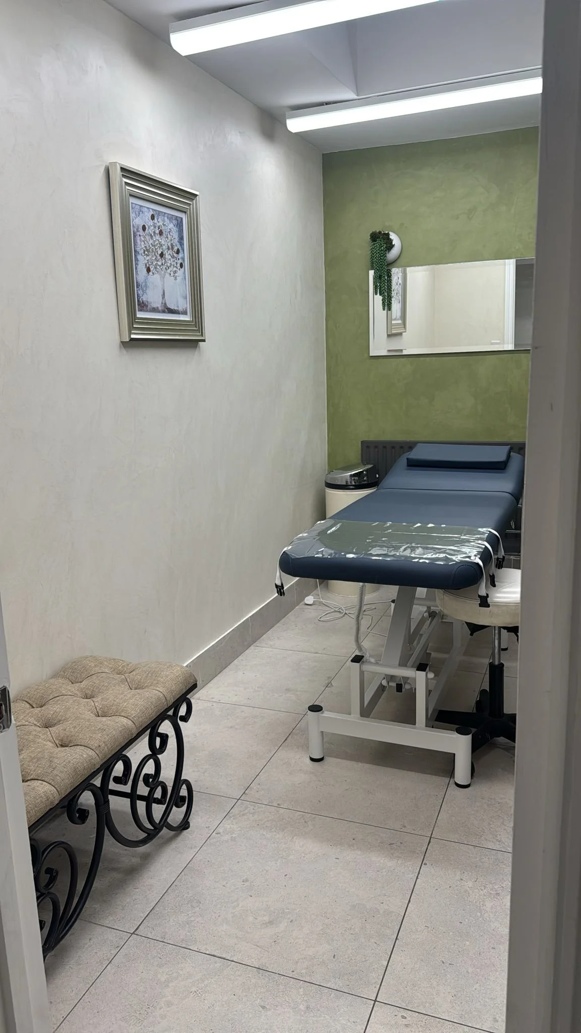 An examination room with a medical examination table, a cushioned bench, wall art, a mirror, and a potted plant, illuminated by fluorescent lighting.