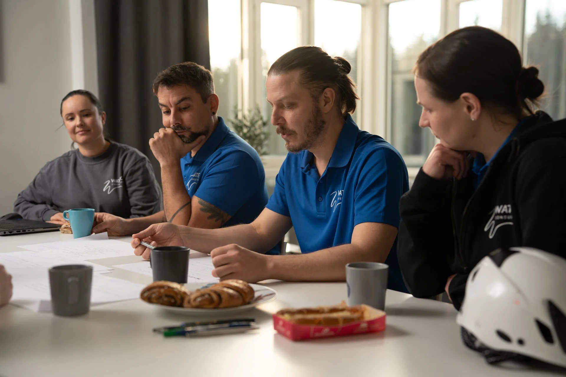 Neljä ihmistä pöydän ääressä, pöydällä mukeja ja kypärä