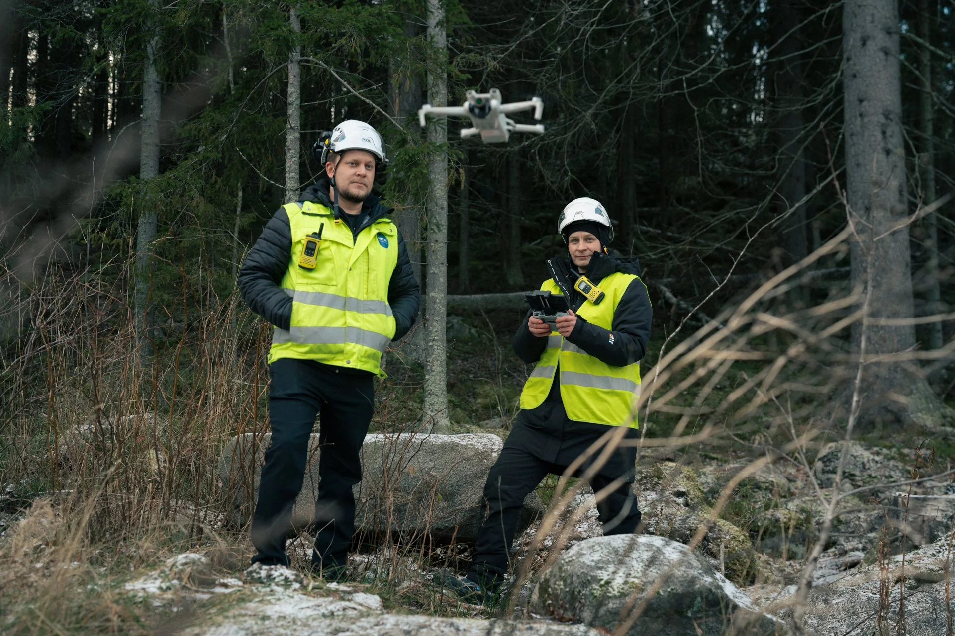 Kaksi työntekijää keltaisissa huomiovaatteissa lennättää dronea.
