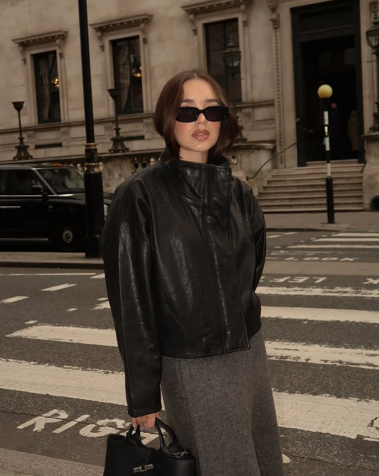 A woman with short brown hair wearing black sunglasses, a black leather jacket, and a gray skirt standing on a city street with historic buildings, parked cars, and a crosswalk in the background.