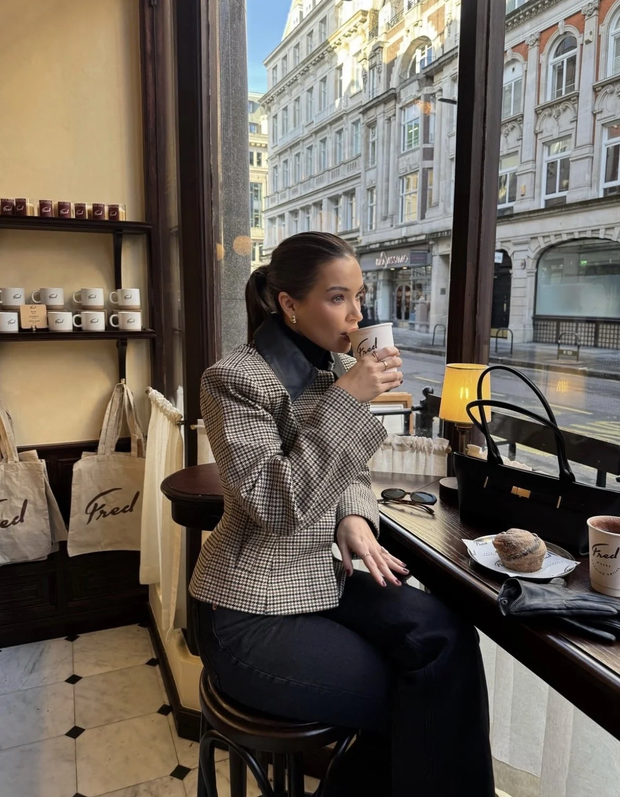 A woman with dark hair in a ponytail, wearing a plaid blazer, sitting at a cafe table near a large window, sipping coffee from a mug labeled 'Fred'. On the table are a cinnamon roll, a pink cup, sunglasses, and a black handbag. Behind her, store shelves display coffee mugs and tote bags labeled 'Fred'. Outside, a city street with historic buildings is visible.