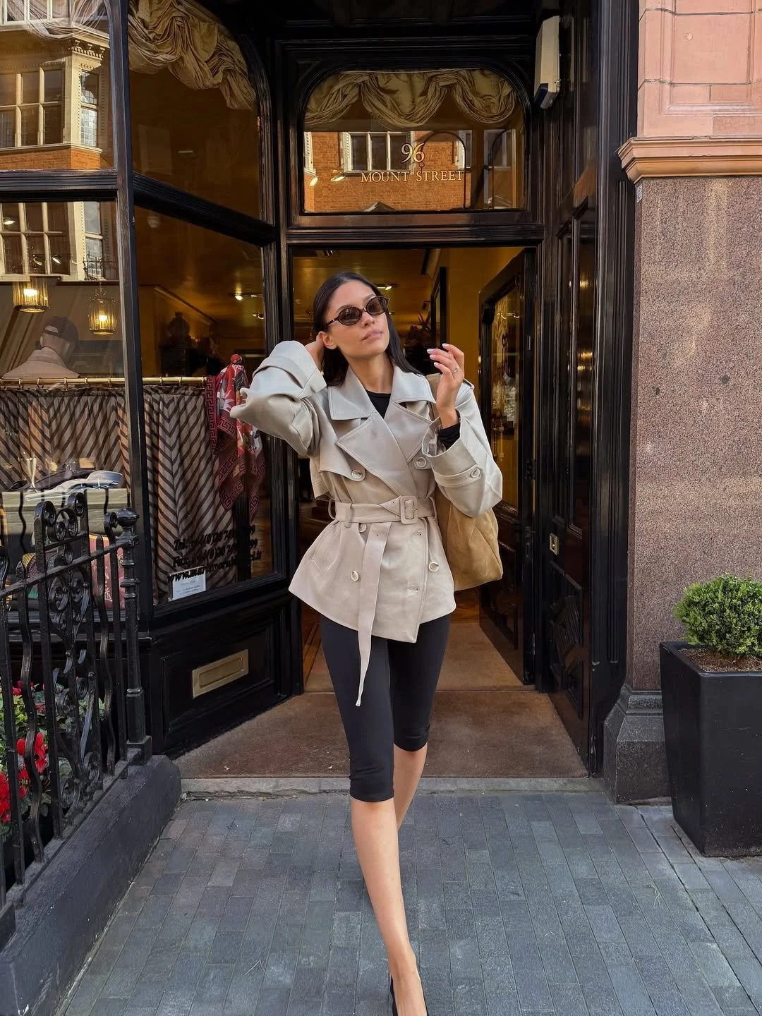 A woman with sunglasses and a trench coat exiting a building on Mount Street, London.