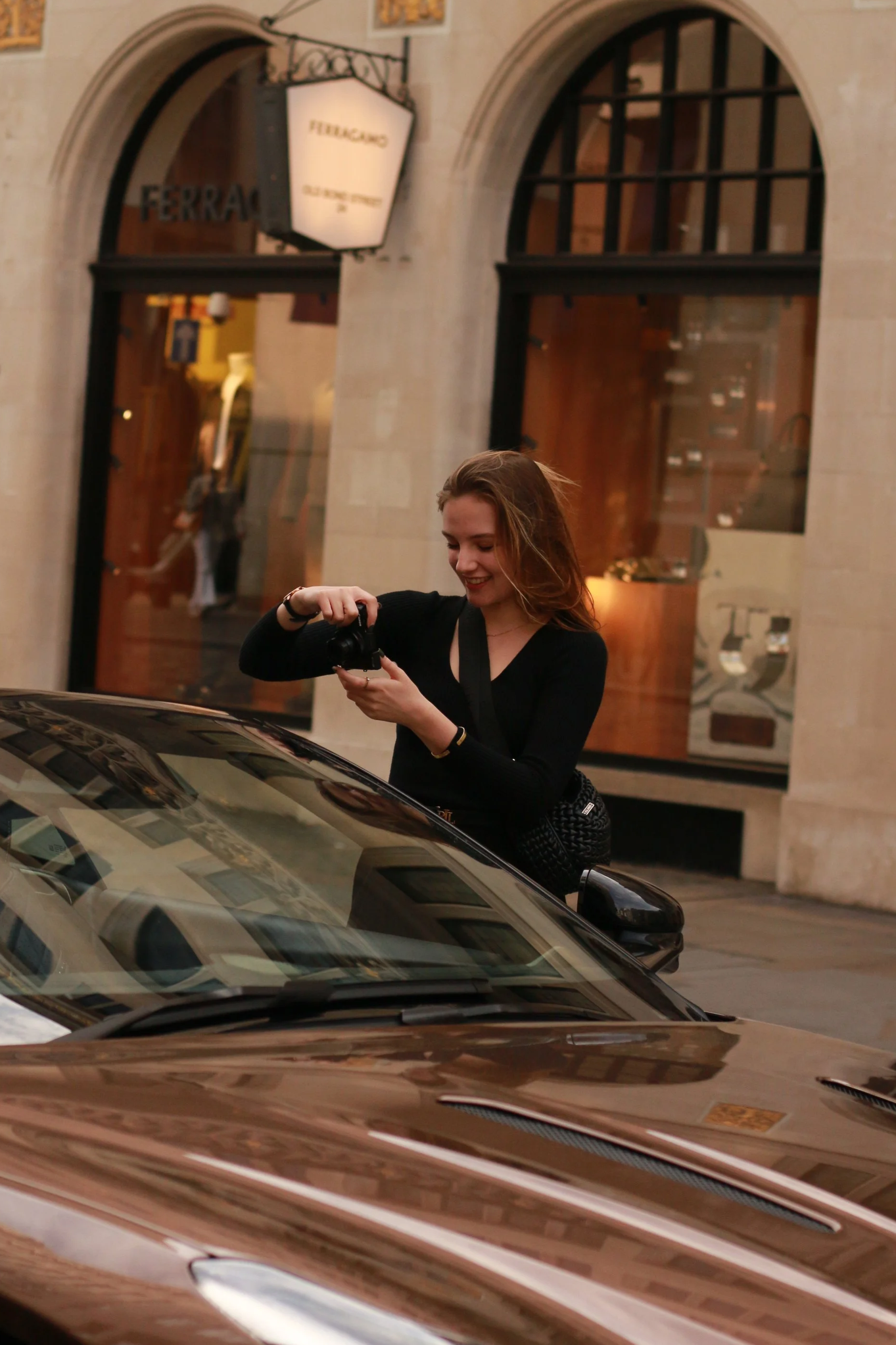 A young woman with long hair smiling and looking at her camera while standing next to a luxury car, with a store sign reading 'FERRAGAMO' in the background.