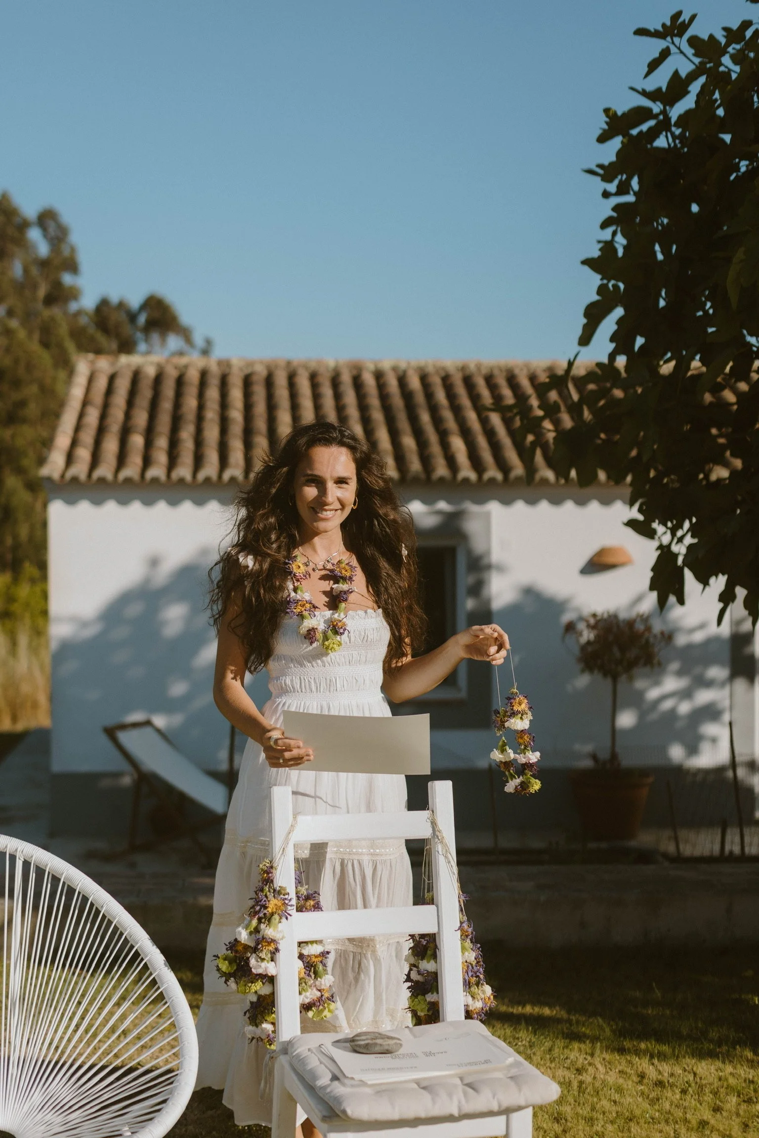 A woman in a white dress with a floral lei stands outdoors in front of a white house with a tile roof. She holds a piece of paper and smiles, standing next to a decorated white chair with flowers. The scene is sunny with green grass, trees, and a clear blue sky.