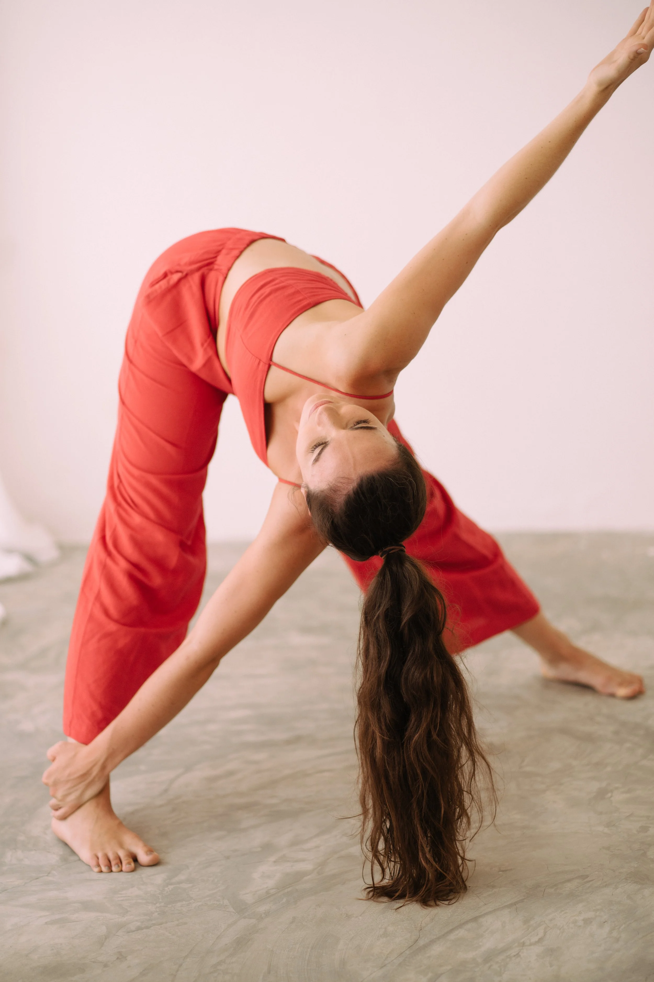 A woman in red yoga attire doing a forward bend stretch with one arm raised and hair hanging down.