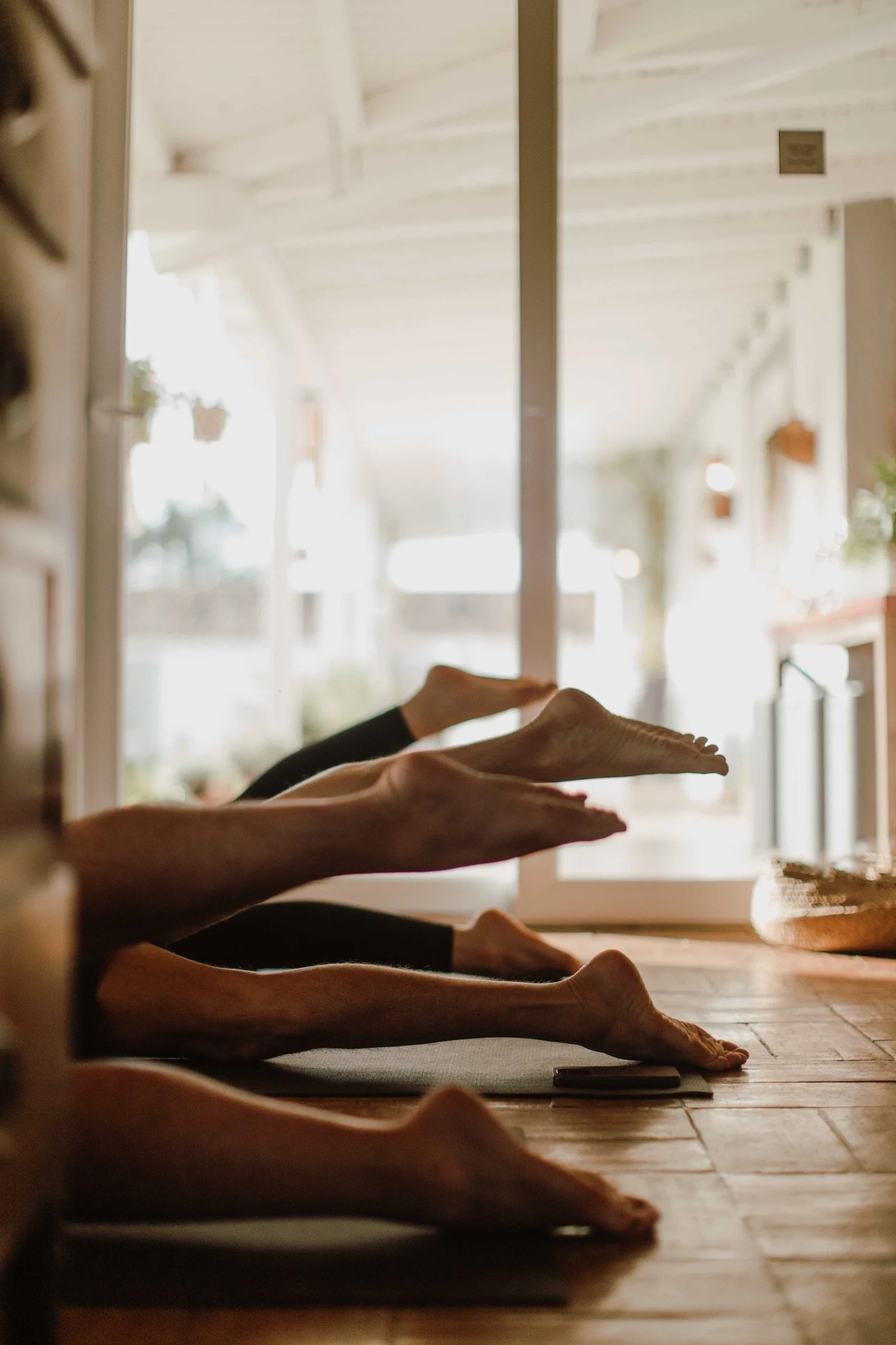 People doing yoga in a row on a wooden floor near a large window, stretching their legs and arms.