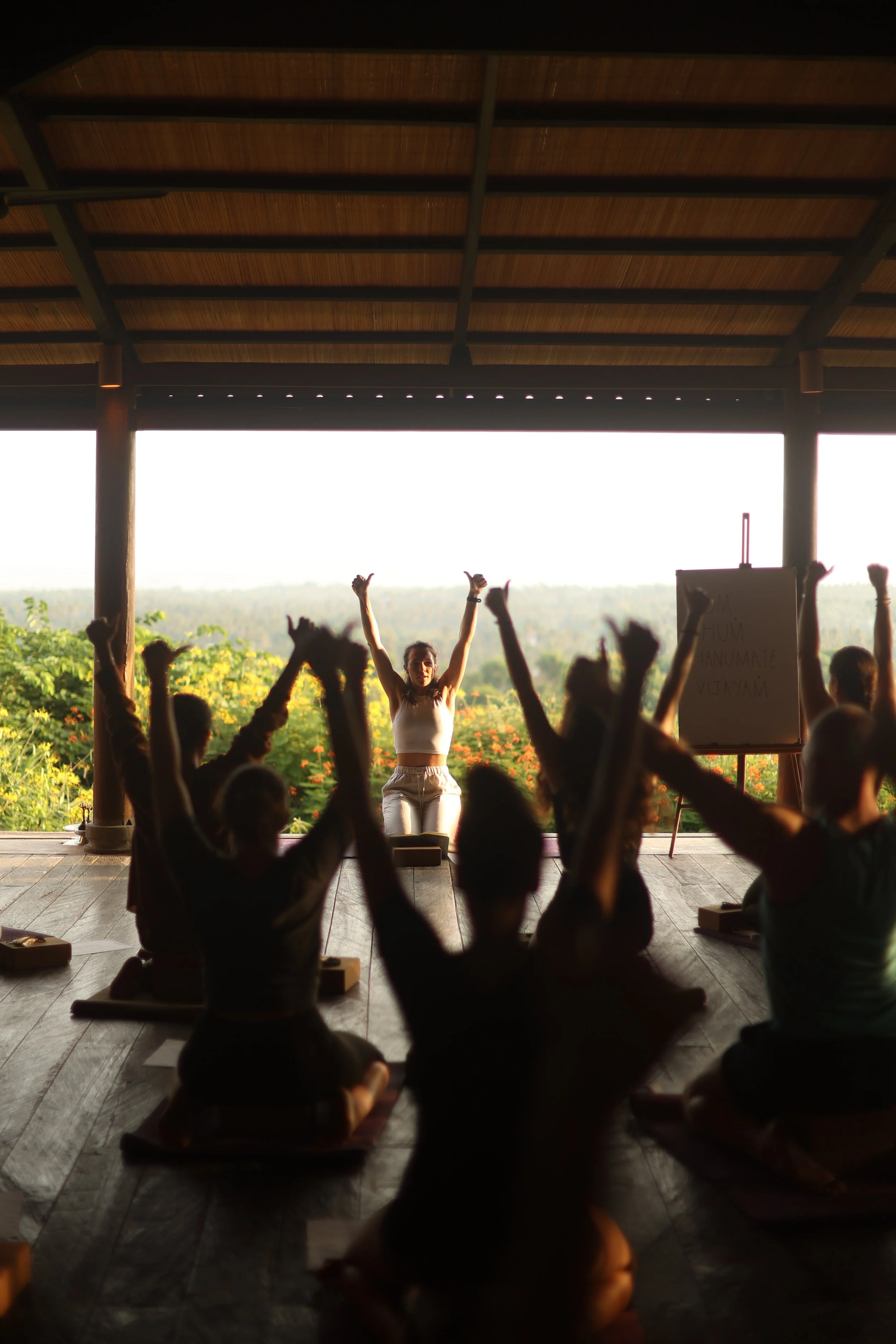 People participating in a yoga or meditation class indoors, with some raising their hands, facing an instructor at the front, with open view of lush greenery outside.