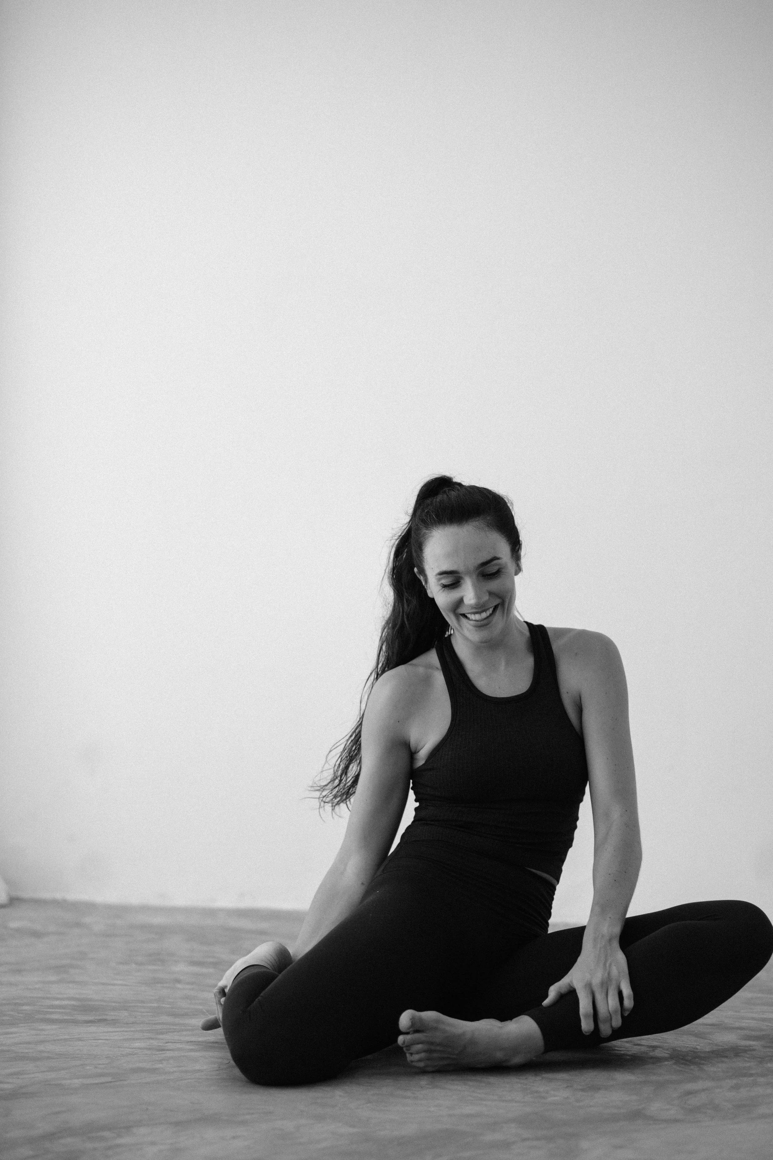 A woman practicing yoga, sitting on a mat in a seated forward bend pose, smiling, in a minimalistic room with a plain wall background.