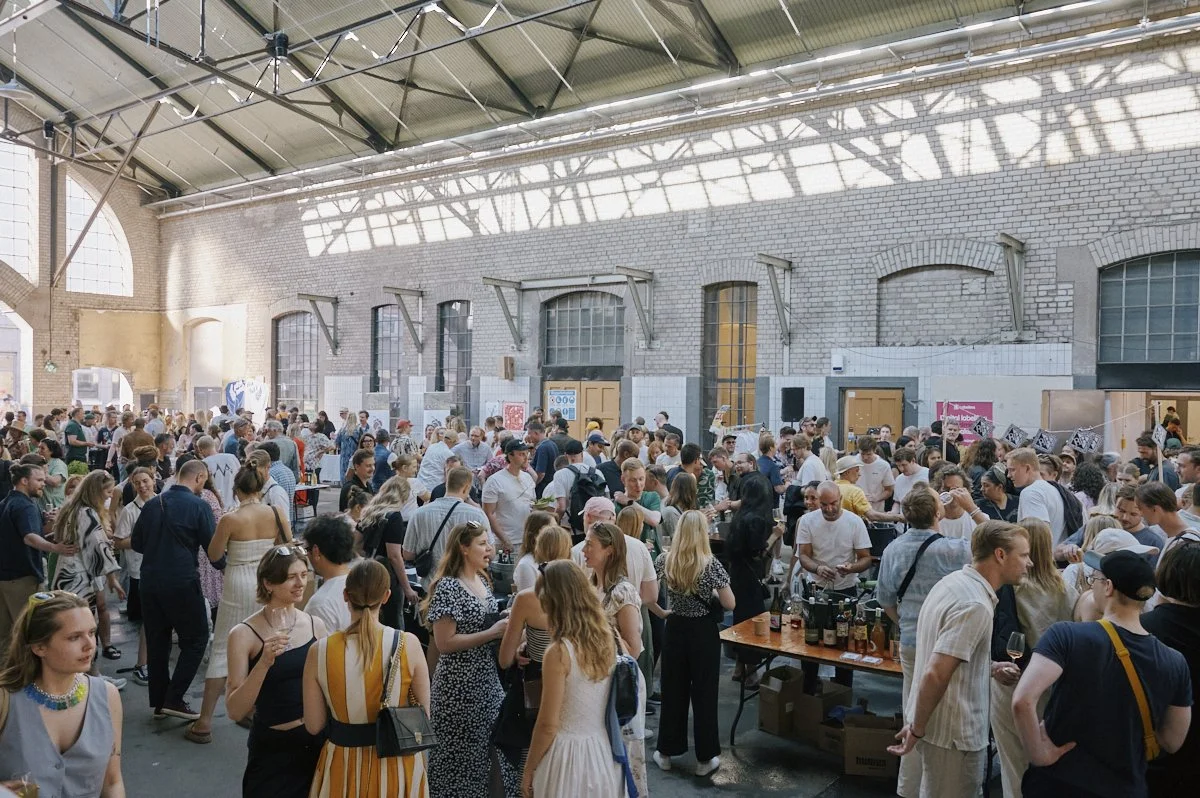 Large crowd of people at an indoor event in a spacious industrial-style venue with high ceilings and brick walls.