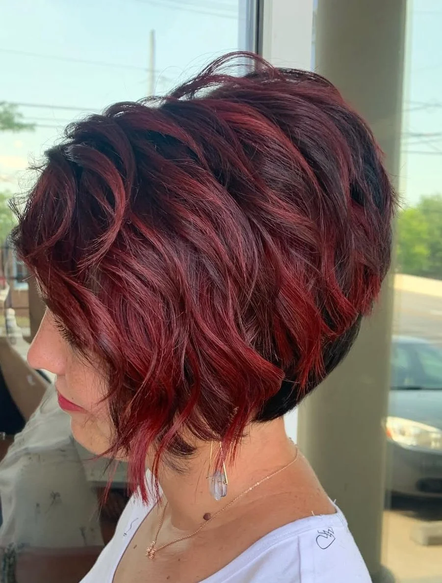 A woman with a short, wavy bob haircut dyed in different shades of red and black, standing indoors near large windows.