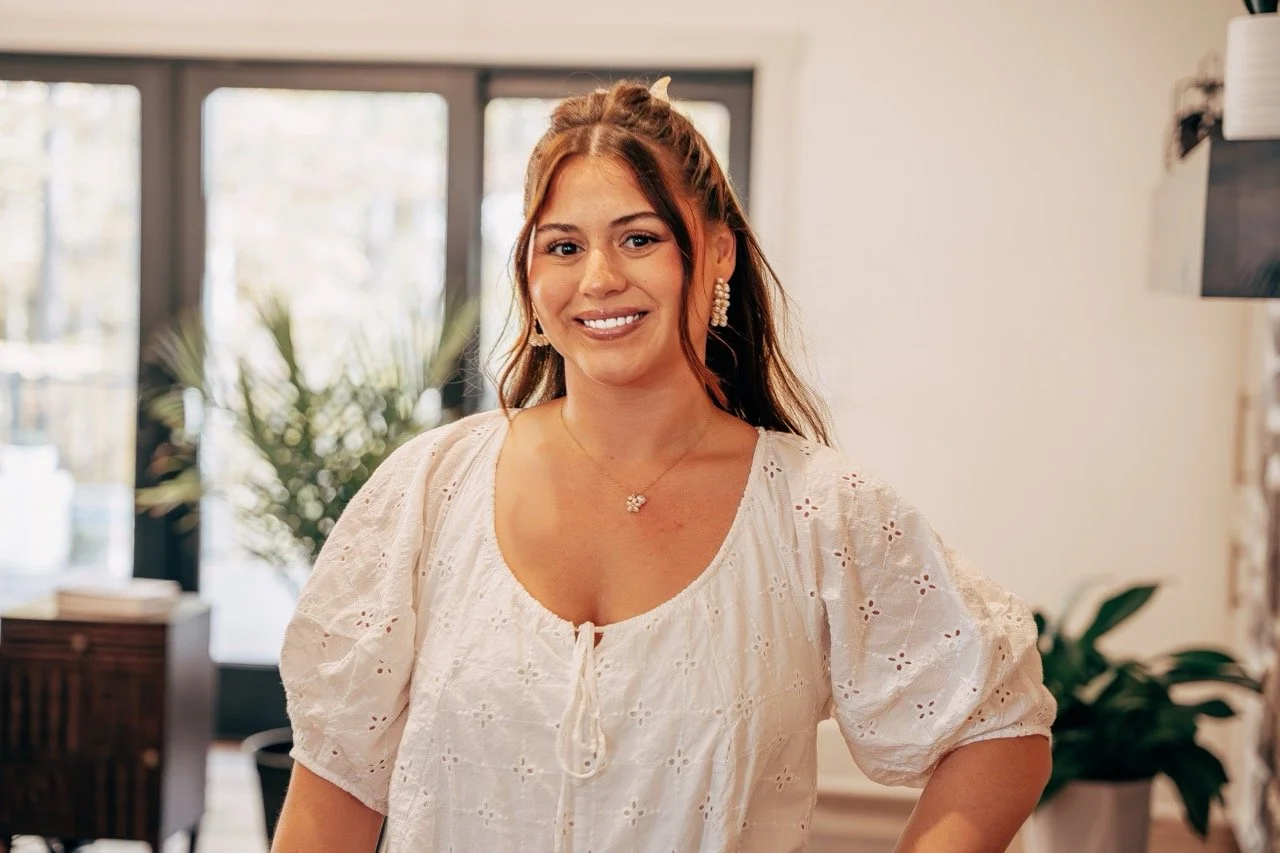 A woman with brown hair wearing a white embroidered blouse and jewelry, smiling indoors with large windows and plants in the background.