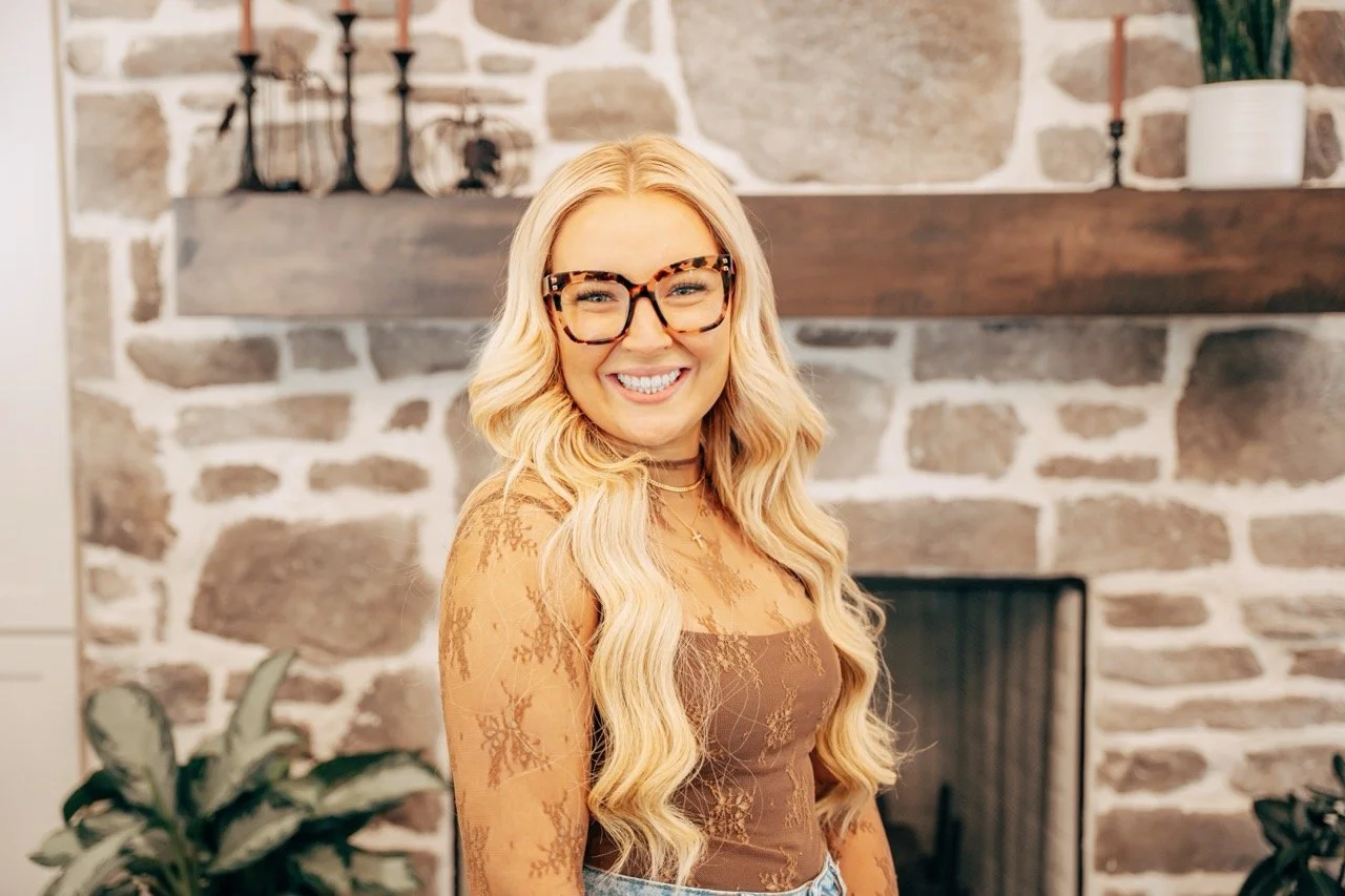 A woman with long wavy blonde hair wearing large leopard-print glasses and a brown lace top, smiling in front of a stone fireplace with a wooden mantel and candles.