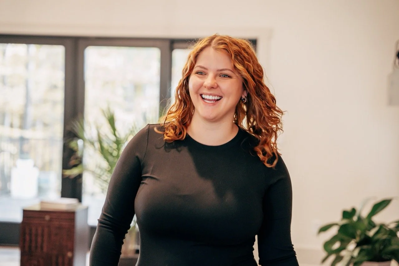 Smiling woman with curly red hair wearing a black long-sleeve shirt standing indoors near a window with plants and furniture in the background.