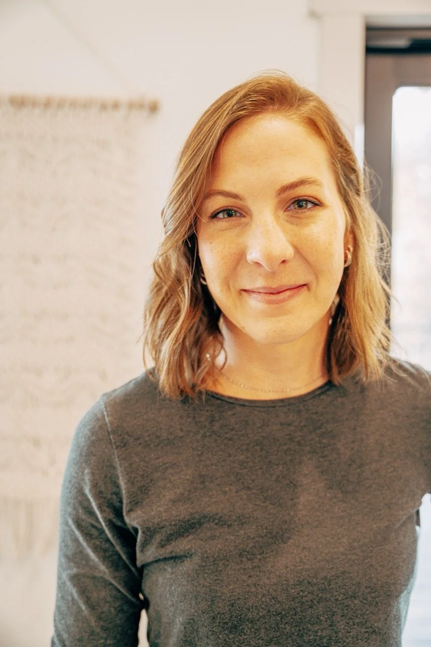 A young woman with wavy, shoulder-length blonde hair, wearing a dark gray top, standing indoors near a window with a bright, blurred background.