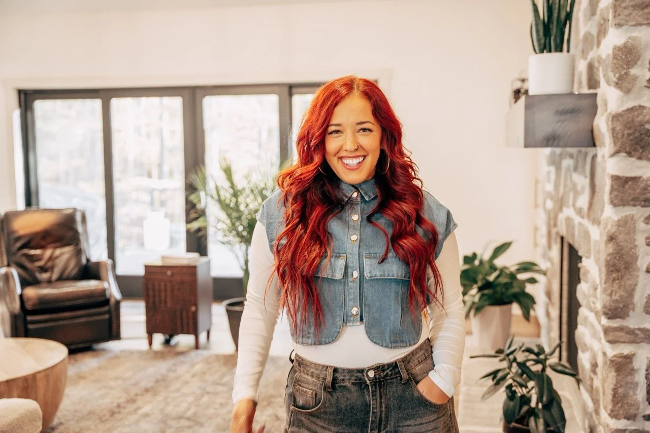 A woman with long red hair, smiling, wearing a denim vest over a white long sleeve shirt, with her left hand in her pocket, standing in a cozy living room with stone fireplace and large windows.