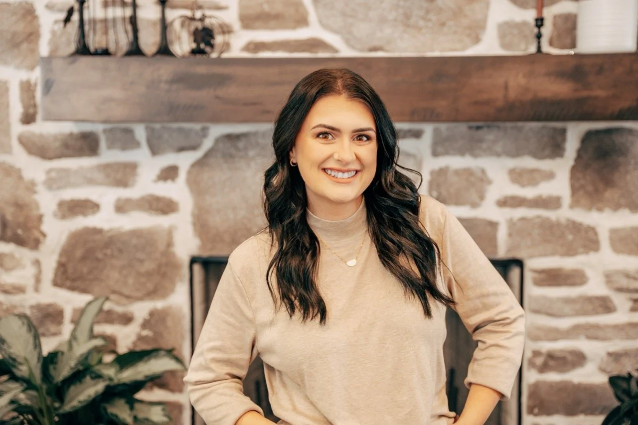 A young woman with long dark wavy hair, wearing a beige top, smiling, standing in front of a stone fireplace with a wooden mantle and decorative items.