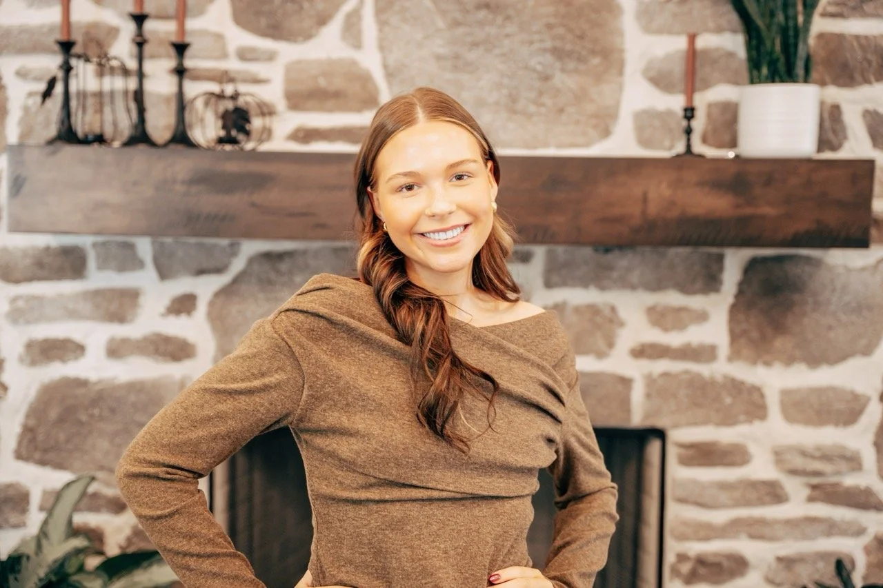 Young woman with long brown hair smiling, standing in front of a stone fireplace with a wooden mantel, decorated with candle holders and potted plants.
