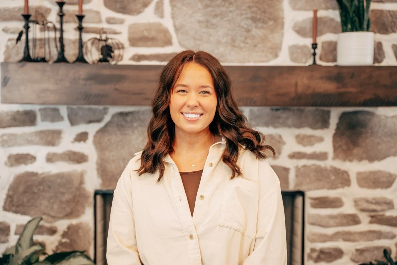 Smiling woman with wavy brown hair standing in front of a stone fireplace with a wooden mantle decorated with black candleholders and small pumpkins.
