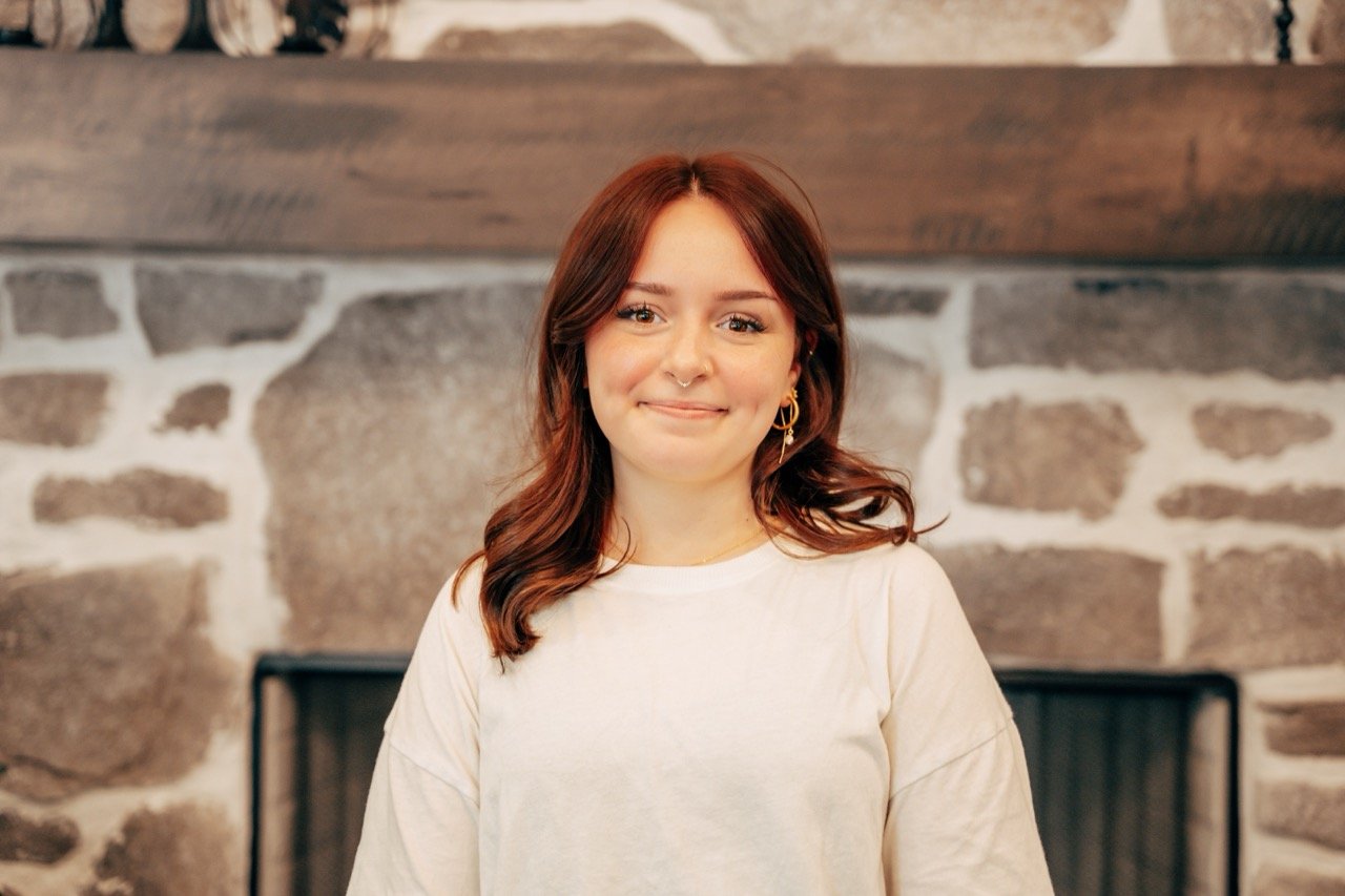 A young woman with shoulder-length brown hair and earrings, smiling slightly, standing in front of a stone fireplace with a wooden mantle.