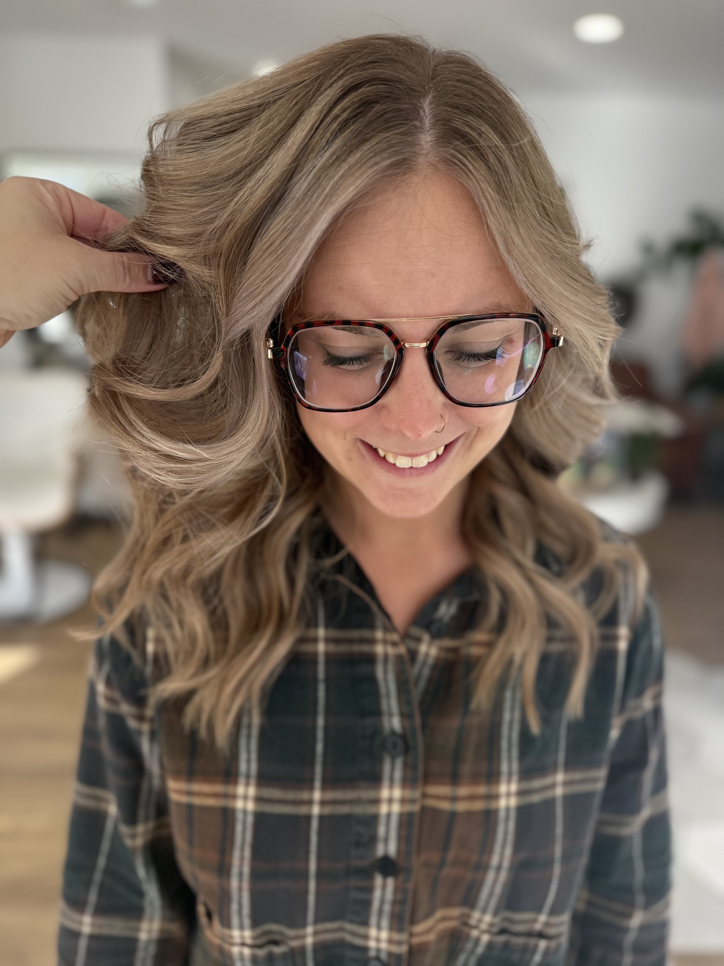 Young woman with wavy blonde hair, wearing glasses and a nose ring, smiling with eyes closed, in a plaid shirt, in an indoor setting.