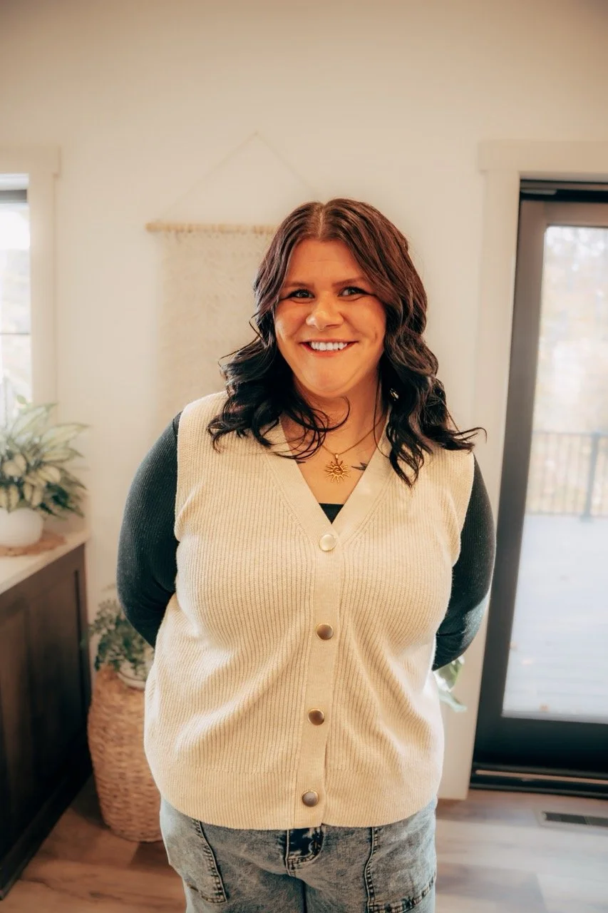 A woman with shoulder-length curly dark hair smiling at the camera standing indoors. She is wearing a cream-colored knitted vest with large buttons over a black long-sleeve shirt and light jeans. There are windows and a door with a deck outside behind her, and indoor plants near a cabinet.
