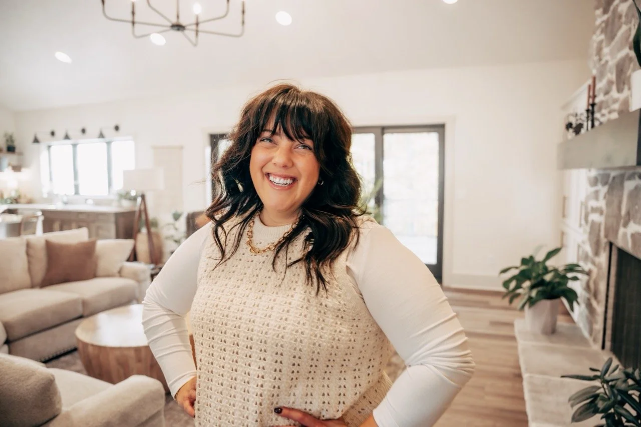 Smiling woman with dark curly hair in a living room with beige couches, a round wooden coffee table, and a stone fireplace.