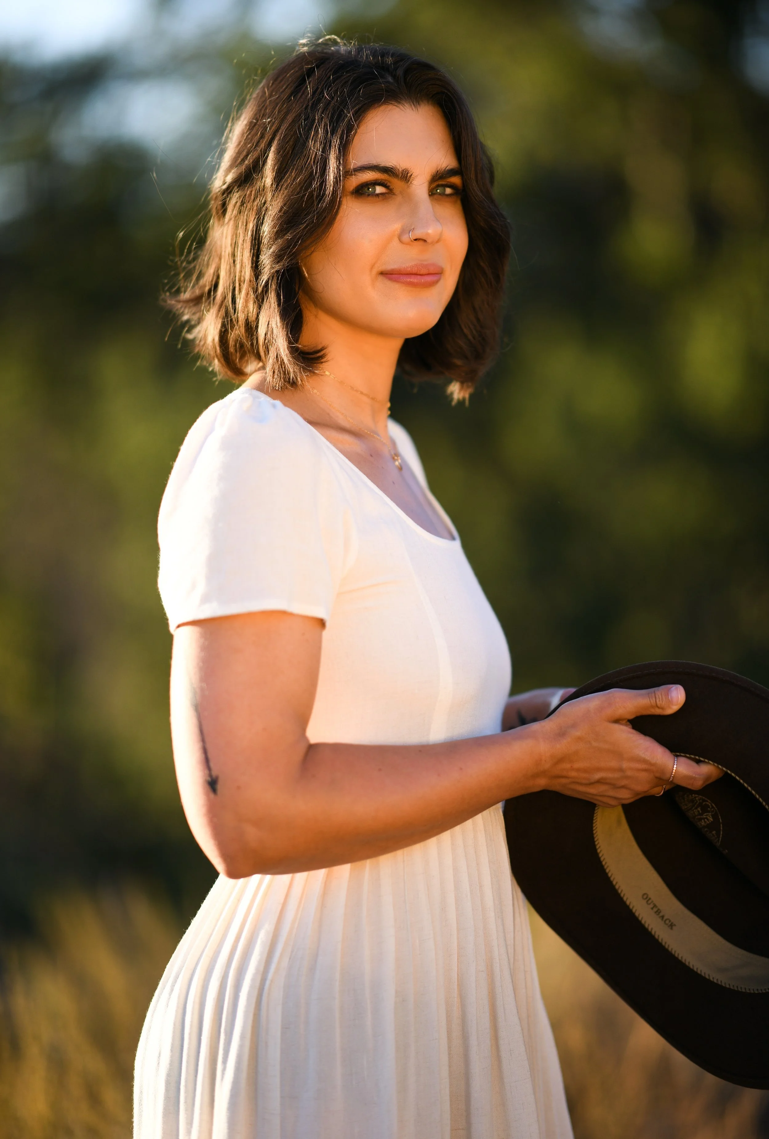 A woman with dark, shoulder-length hair, wearing a white dress, holding a black hat, outdoors with blurred green foliage in the background, during sunset.