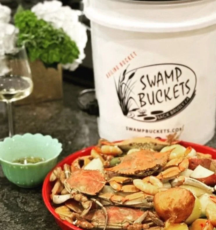 A bucket labeled 'Swamp Buckets' contains cooked seafood, including crabs, shrimp, and potatoes. Nearby, a glass of white wine, a green dipping bowl with sauce, and a vase of flowers are on a dark countertop.