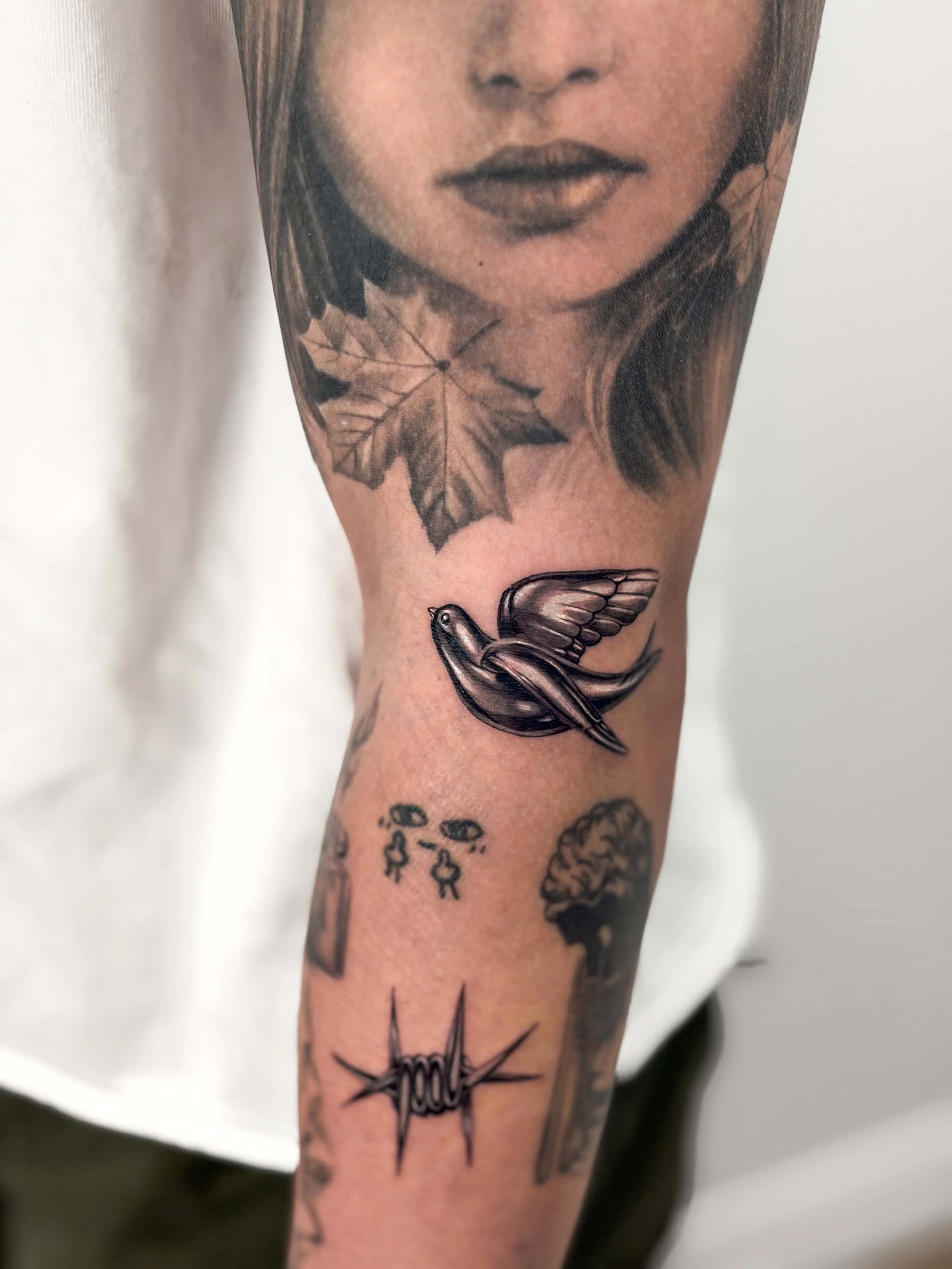 Black and gray tattoo of a woman’s face and leaves on upper arm, with a chrome effect metal silver flying swallow bird, and a barbed wire.