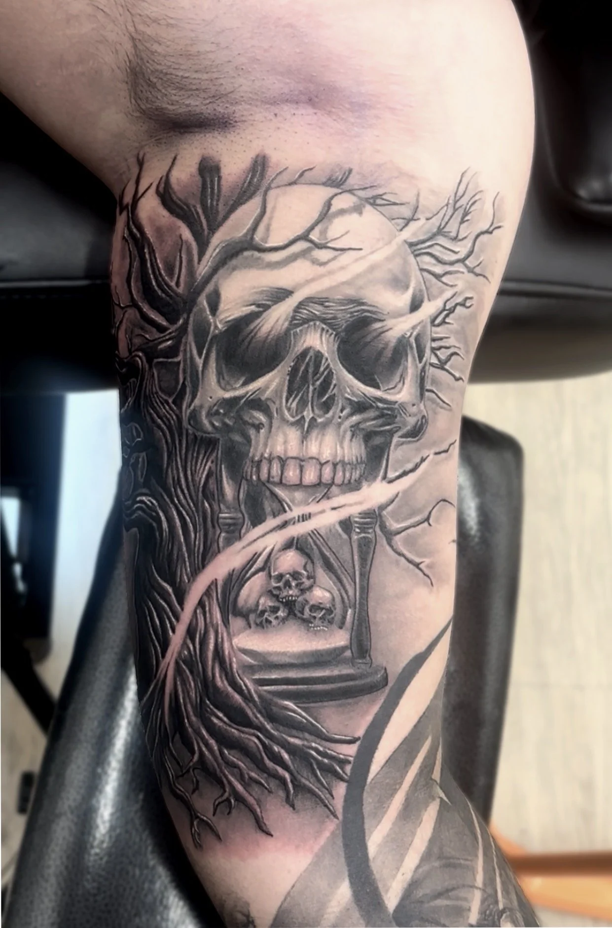 Black and grey realism tattoo of a skull with tree branches, an hourglass, and wind bars on the person's arm.