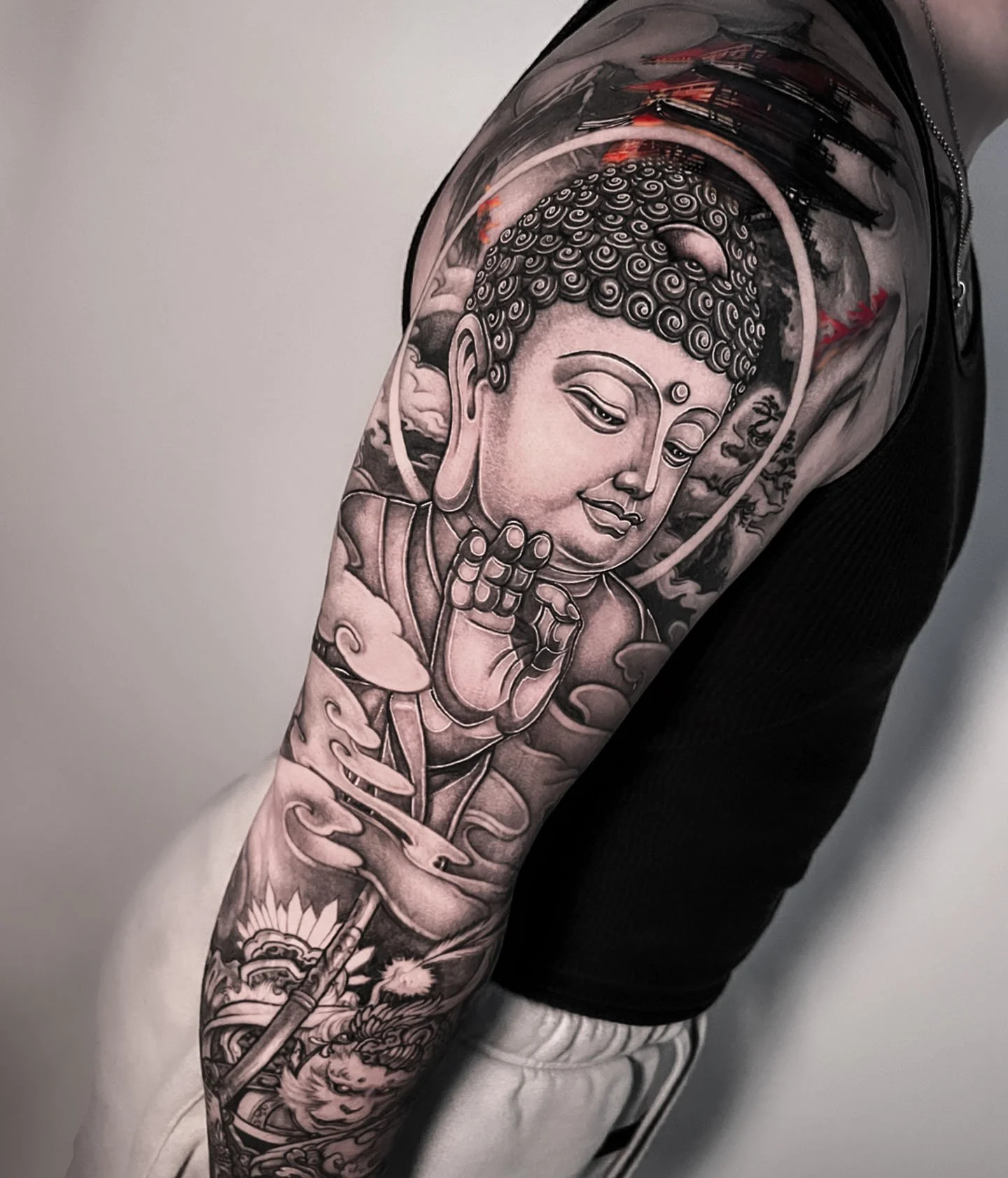 Black and grey Japanese/Asian tattoo of a serene, smiling Buddha with a halo, surrounded by clouds and decorative elements on a person's upper arm.