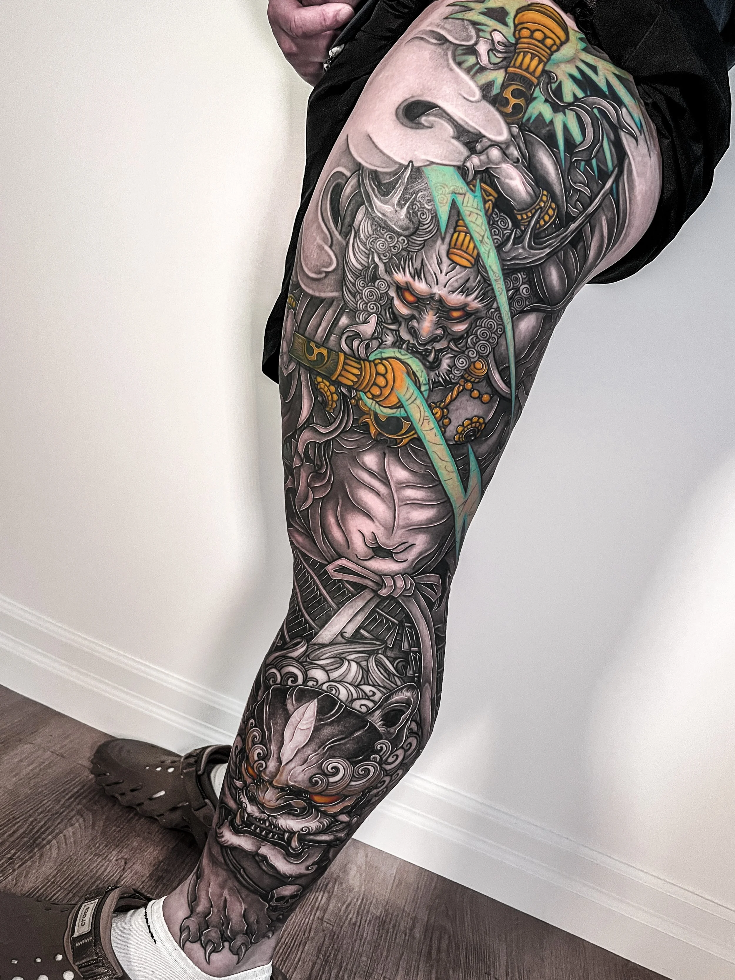 A person's leg sleeve with a colorful Japanese-style tattoo featuring Raijin and a Foo dog, with pops of color, intricate details and shading.