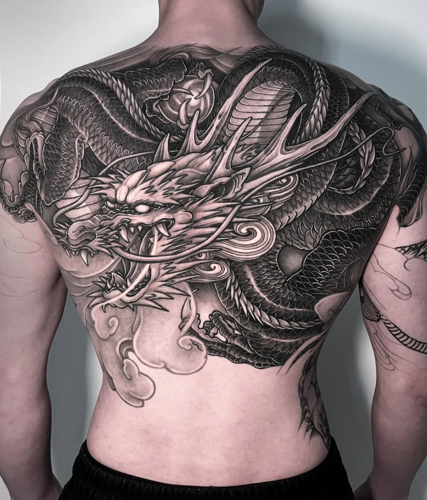 Black and grey tattoo of a fierce dragon on a man's back with detailed scales, horns, and open mouth showing sharp teeth.