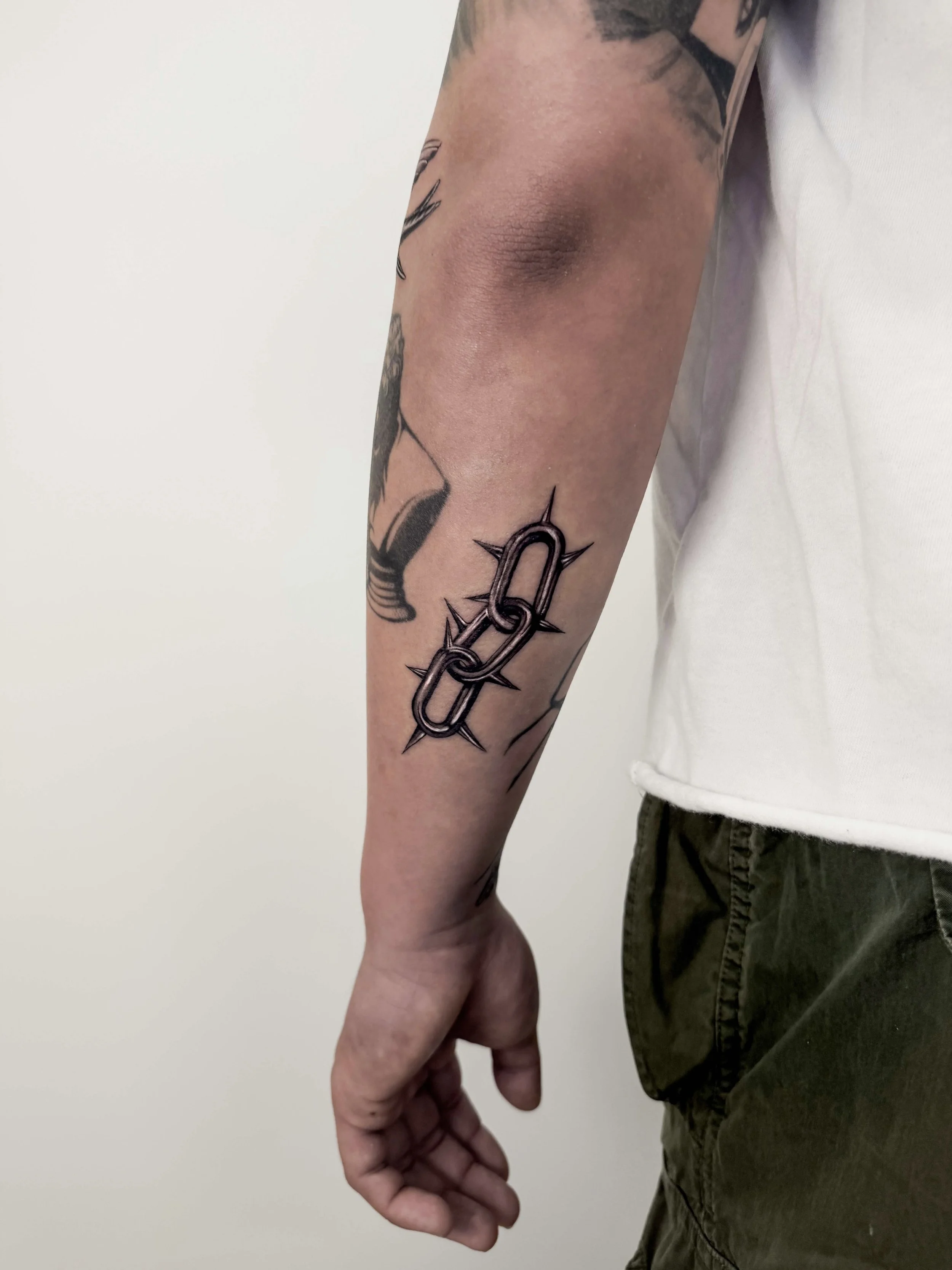 Tattoo of a chrome effect metal silver chain link with spikes on a person's forearm.