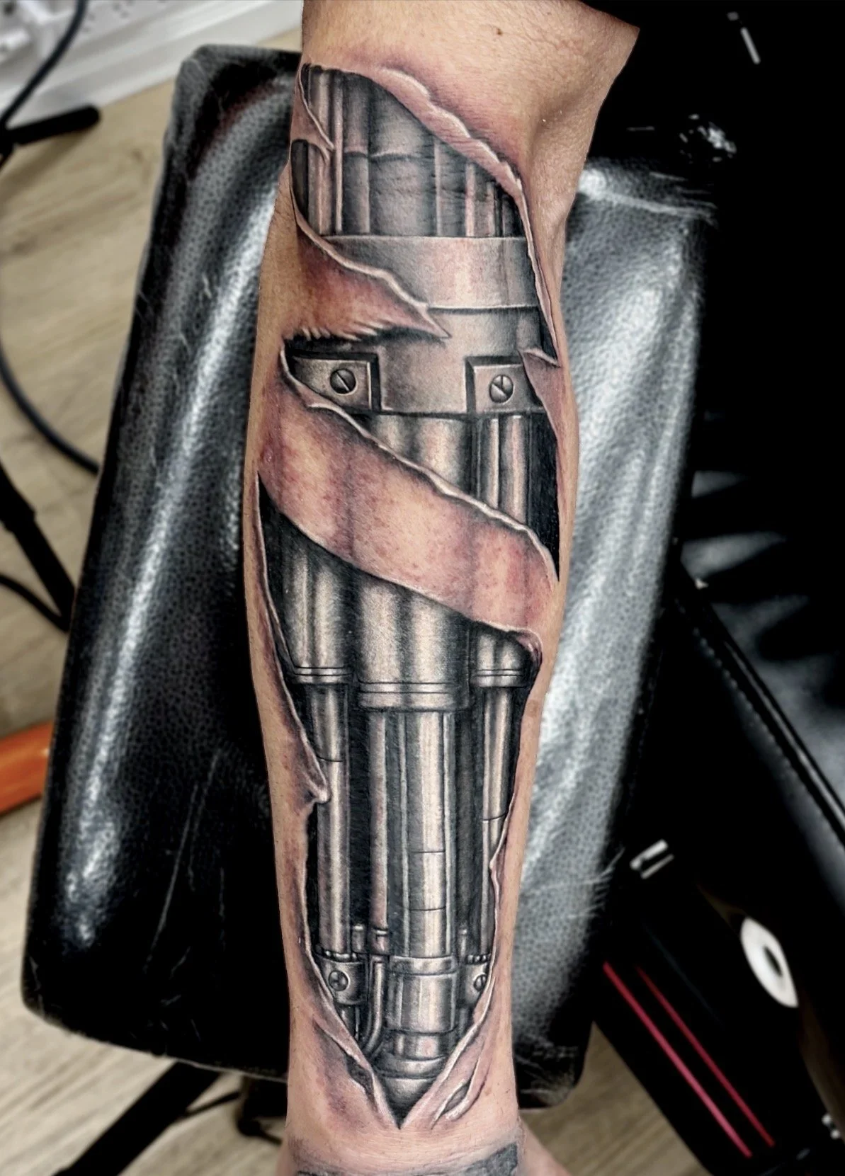 Realistic black and grey realism tattoo of metal pistons appearing as if placed within a person's arm through ripped skin.