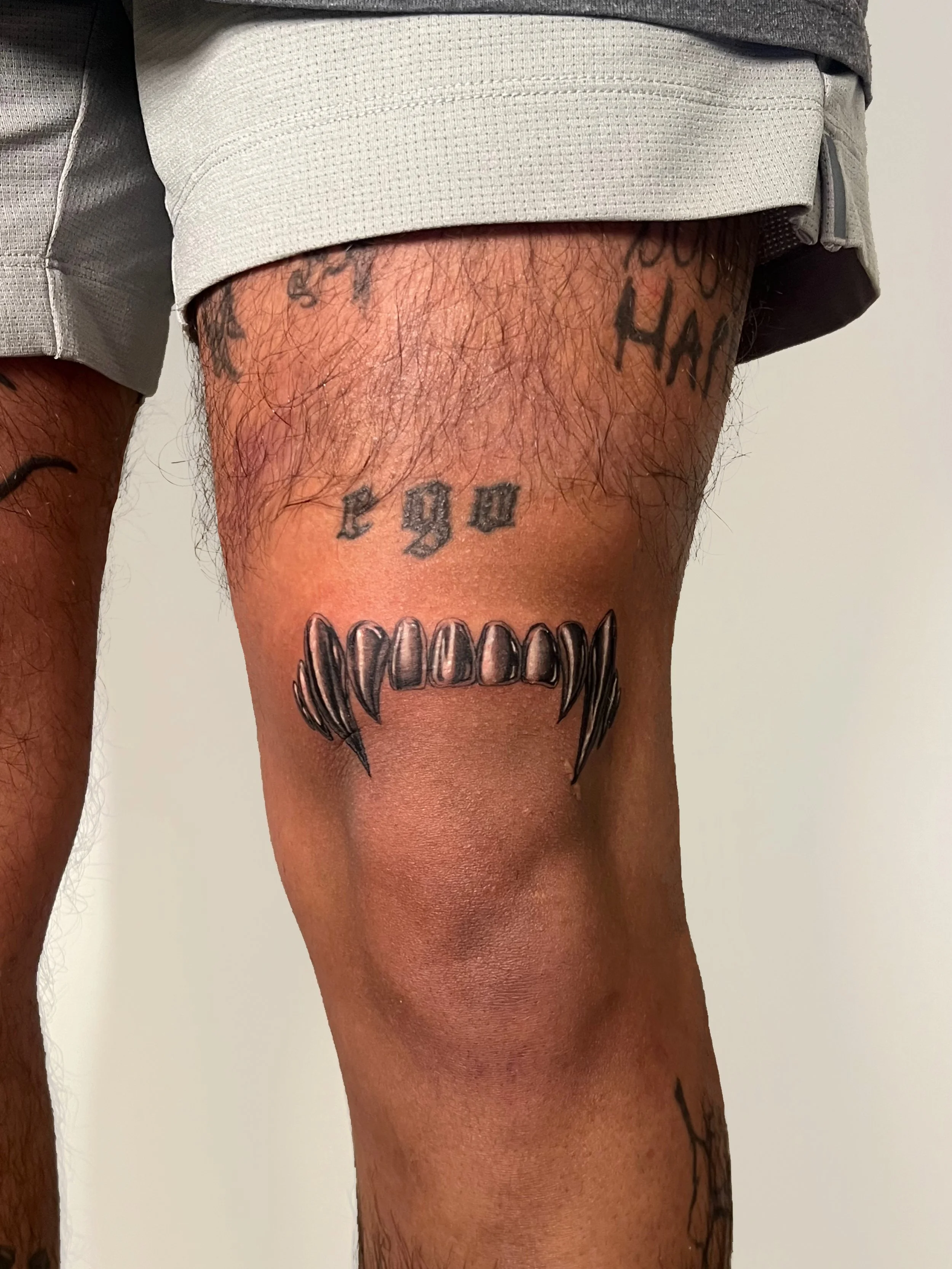 Tattoo of chrome vampire fangs teeth and a tongue on the upper thigh of a person with other tattoos visible on the thigh.