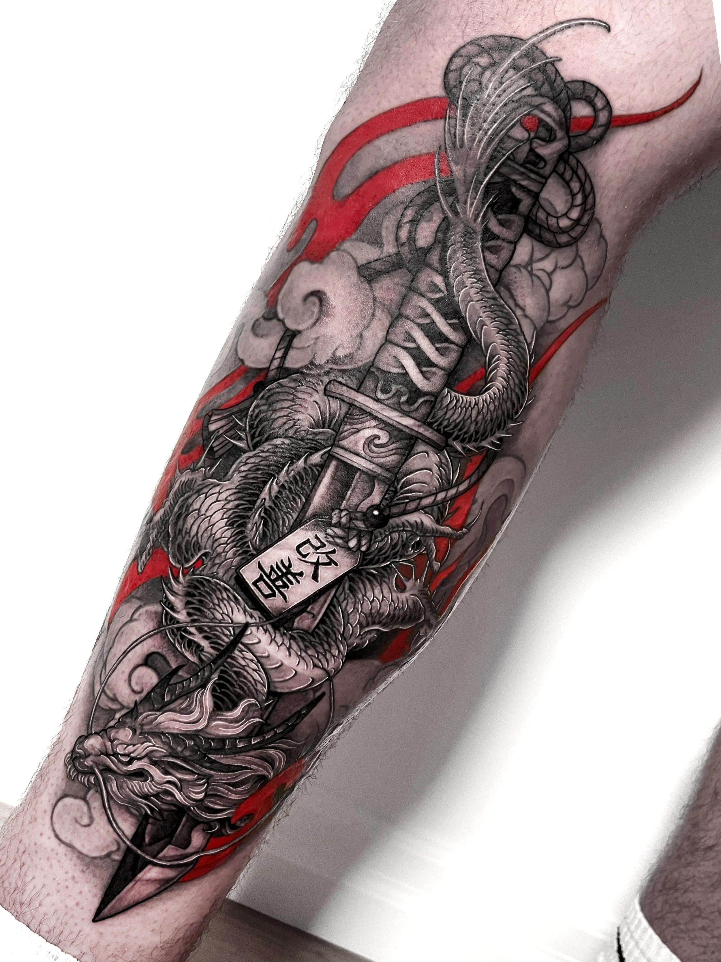 Detailed black and grey Japanese-style dragon tattoo on arm, featuring a sword, a dragon, clouds and red accents.