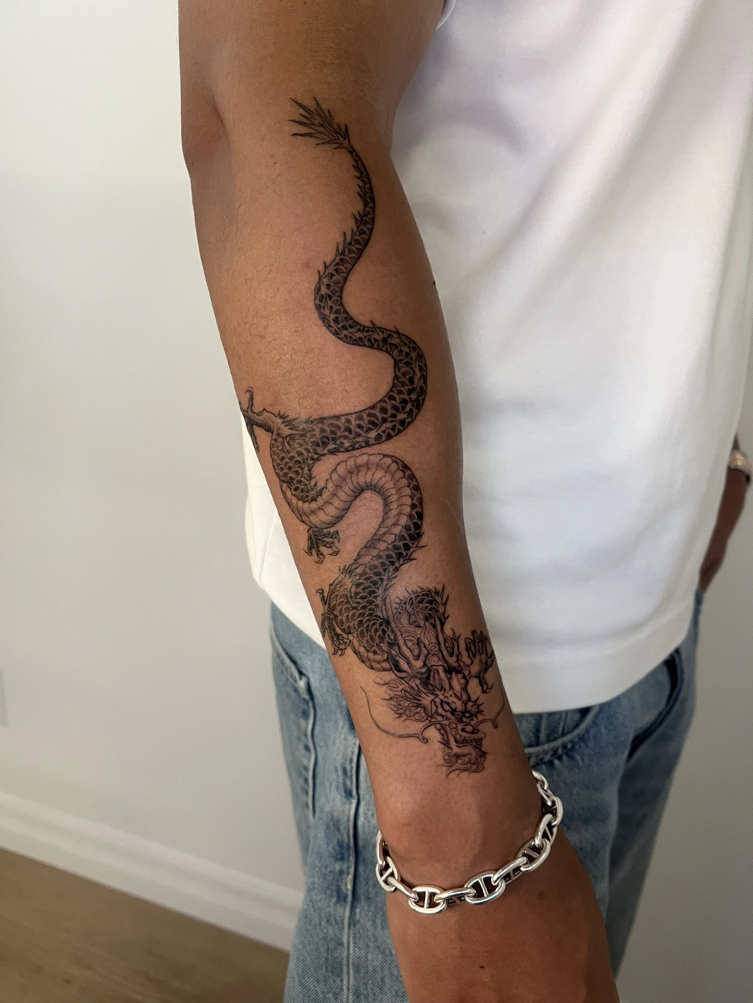 Tattoo of a detailed, traditional Chinese Japanese Asian dragon on a person's forearm, with a silver chain bracelet.