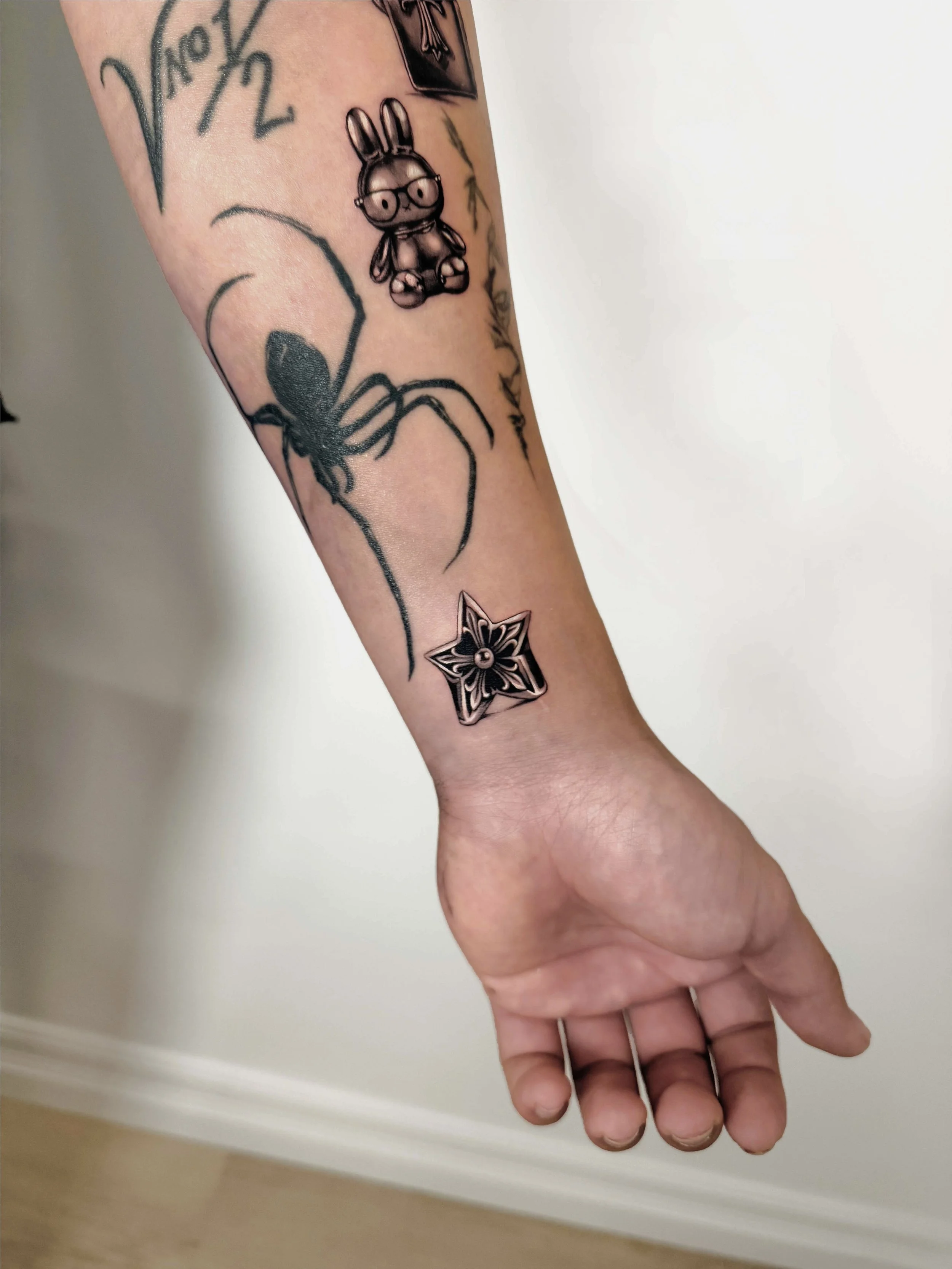 Tattooed arm with various tattoos, including a spider, a bunny Miffy Sanrio character with glasses, and a star-shaped chrome hearts ring signet style silver design.