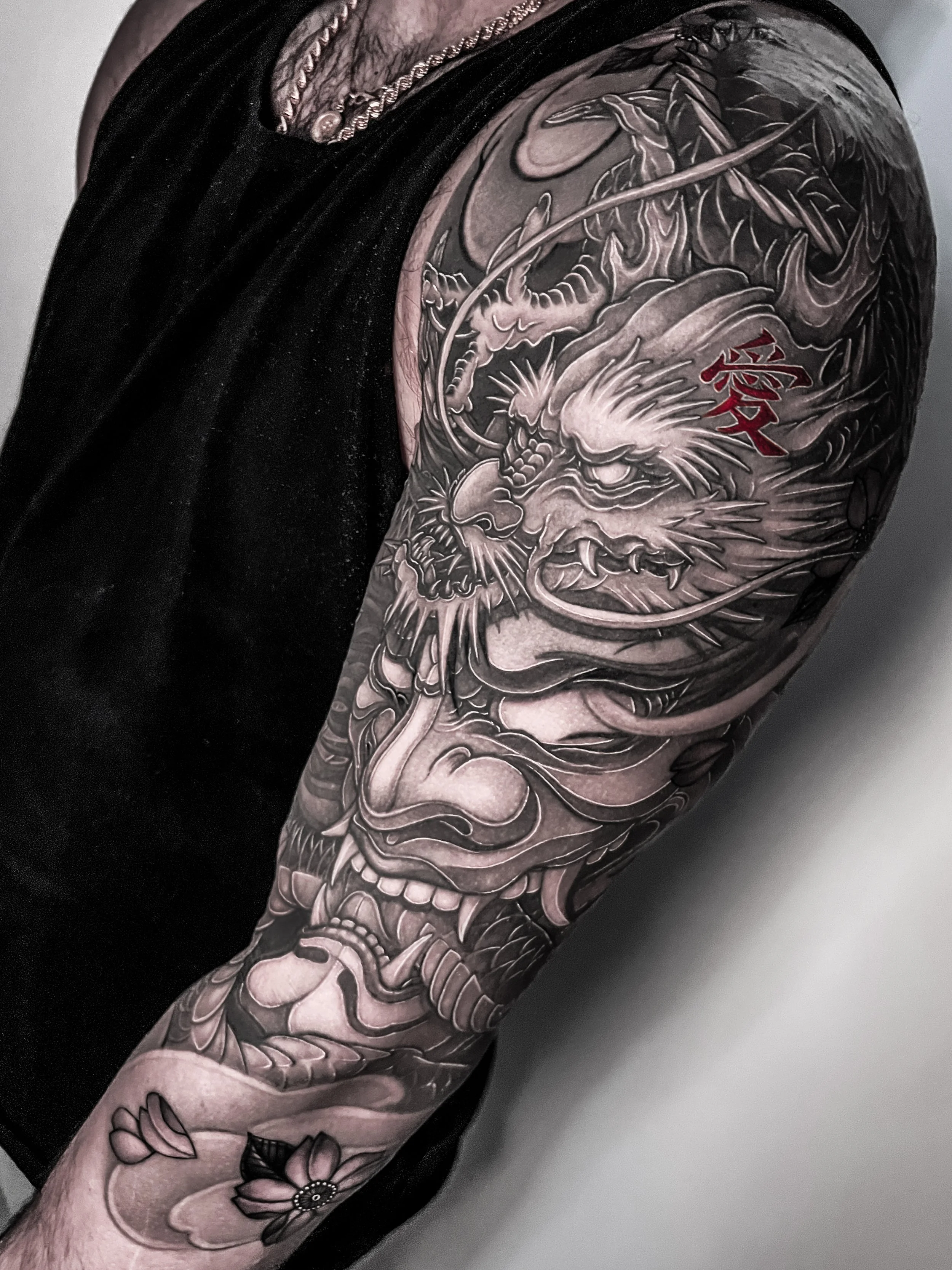 A detailed Japanese/Asian black and grey tattoo of a fierce dragon's face with sharp features and flowing scales on a person's upper arm and shoulder. There is a Hannya Mask, red Japanese characters and a small lotus flower on the forearm.