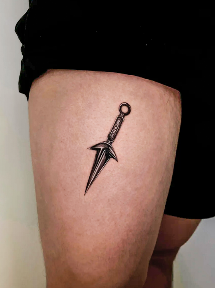 Black and grey realistic tattoo of a Japanese kunai on a person's upper thigh.