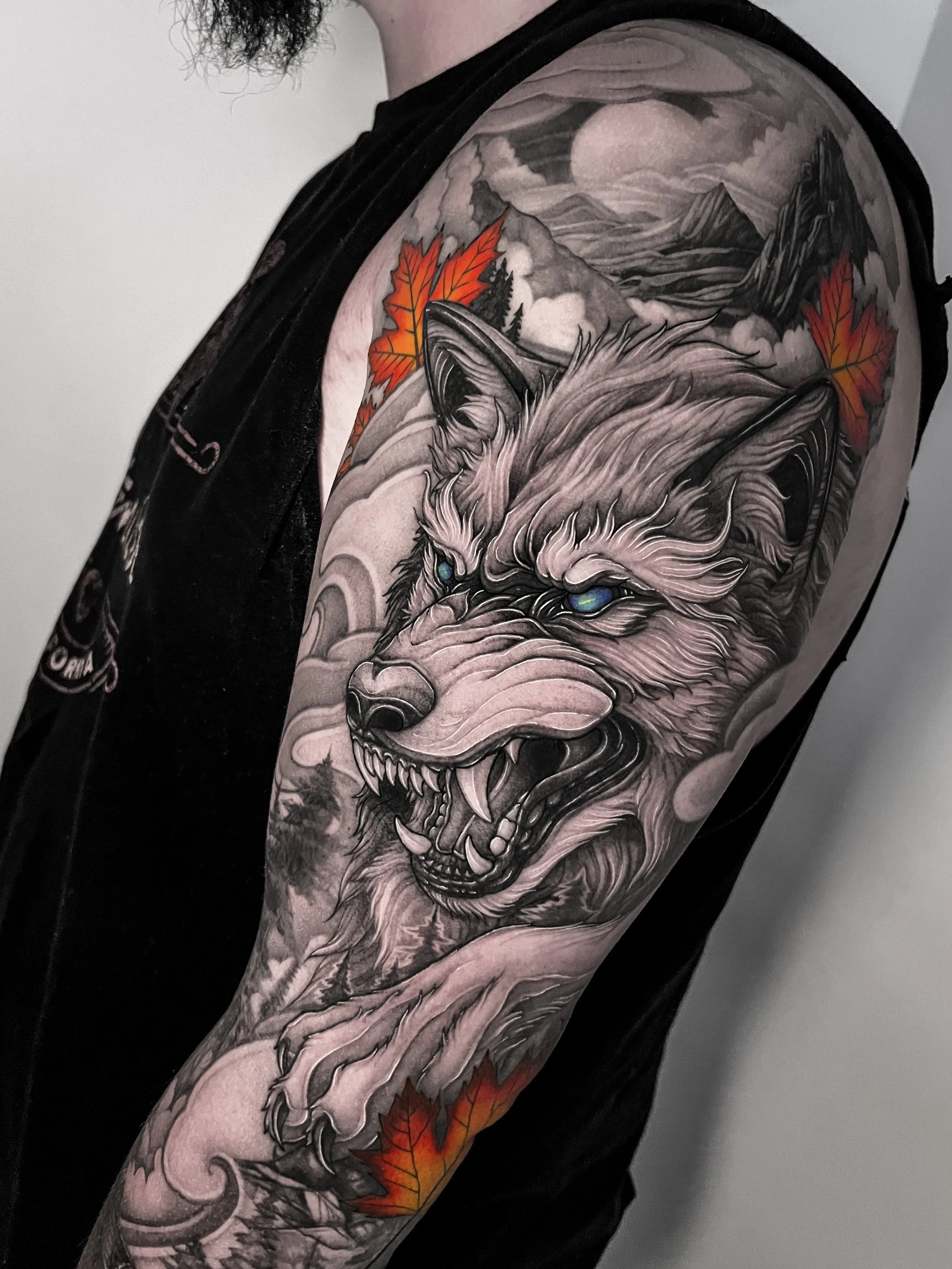 Black and grey tattoo of a snarling wolf with blue eyes on a person's upper arm, with mountains, a full moon, and colorful autumn leaves in the background.